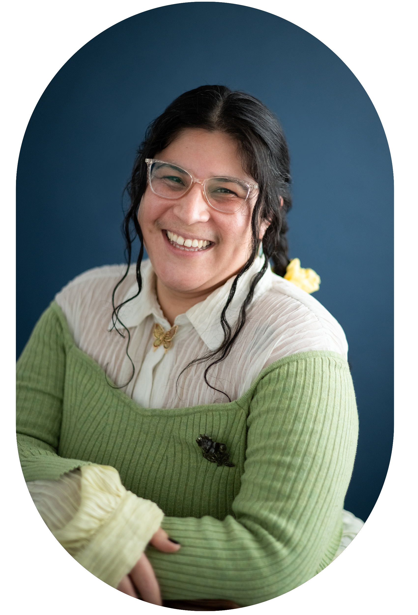 A person with glasses and dark hair styled in braids, smiling, wearing a white blouse with a butterfly brooch and a green sweater with a bee pin, against a blue background.