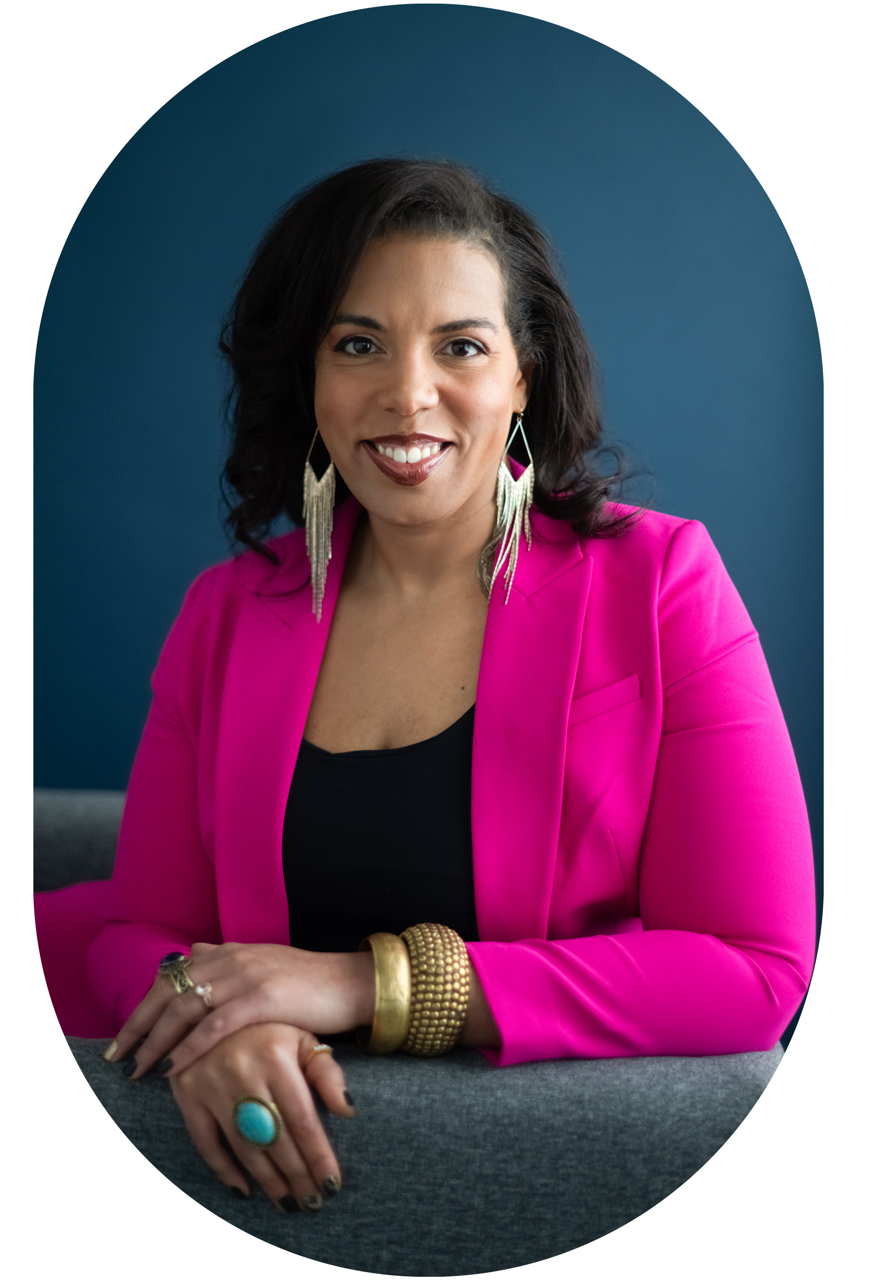 Person wearing a pink blazer and black top, sitting on a gray chair with their hands resting on the armrest, smiling at the camera with long earrings and colorful jewelry, against a dark blue background.