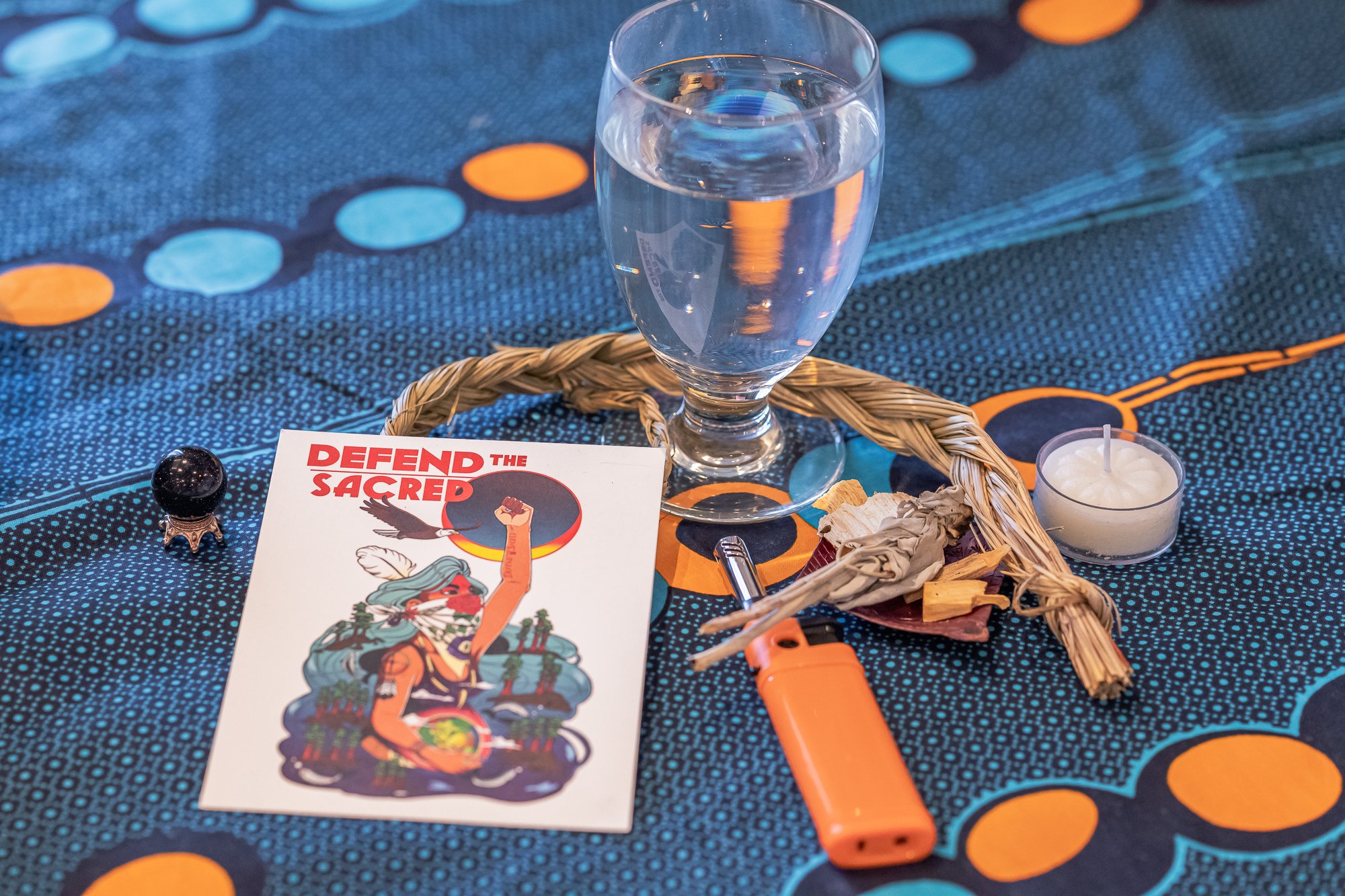 A table with a blue patterned tablecloth, a glass of water, a small candle, a crumpled paper, orange lighter, a tarot card with a person holding a raised fist, and small objects including a black decorative item and a piece of straw.