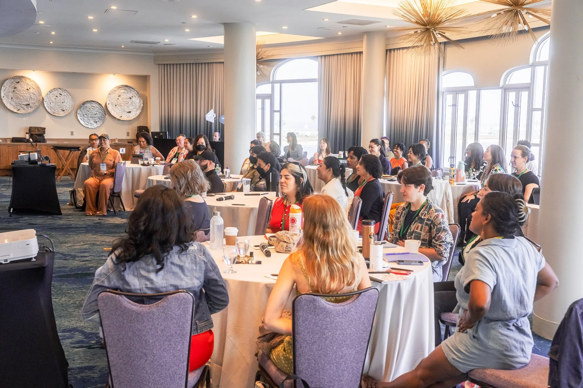 A diverse group of people sitting at round tables during a conference or seminar, listening attentively, with some taking notes and others smiling. The room has large windows, curtains, and decorative wall art.