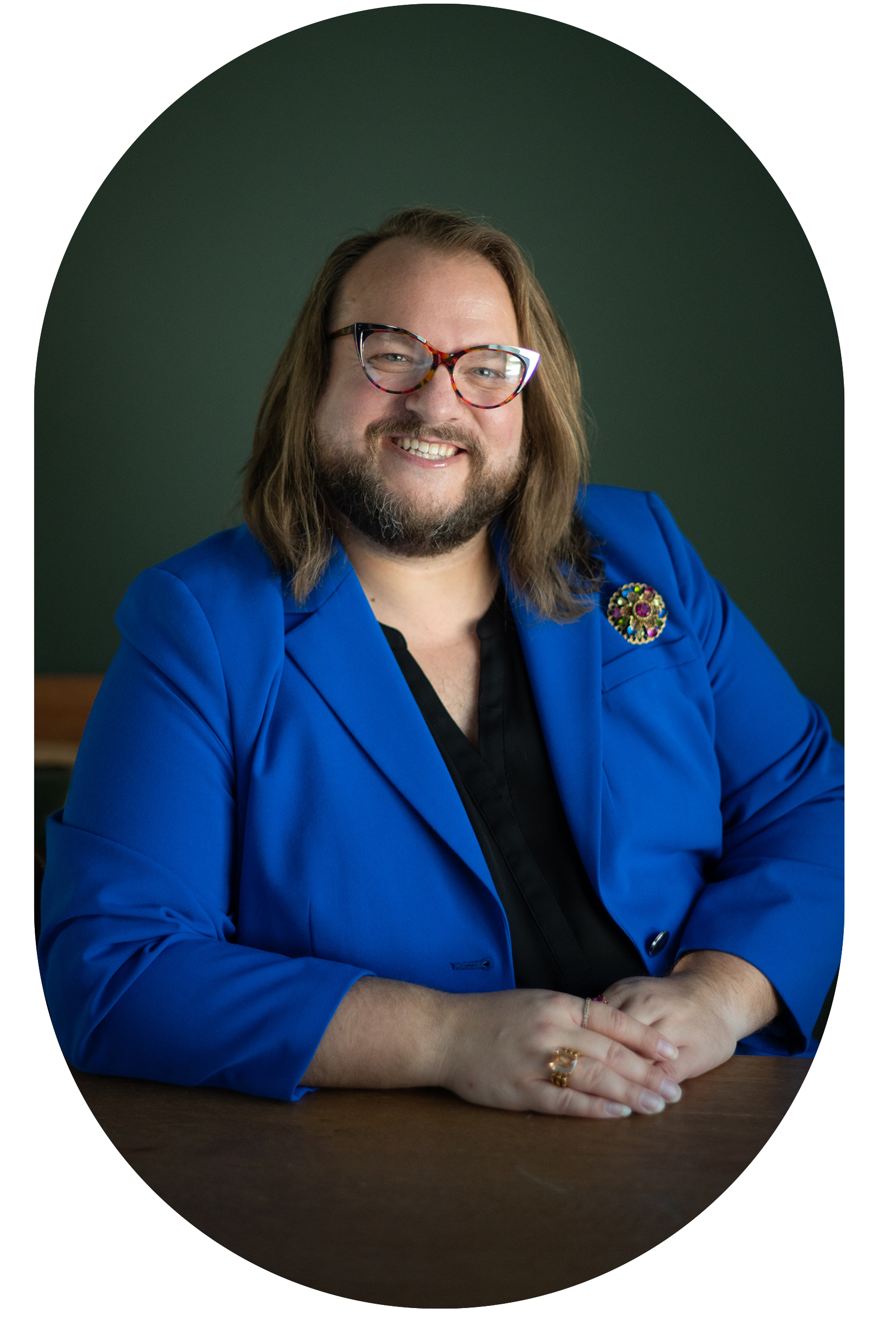 A person with long hair, glasses, and a beard, smiling while sitting at a wooden table. They are wearing a bright blue blazer with a colorful pin on the lapel and a black shirt underneath.