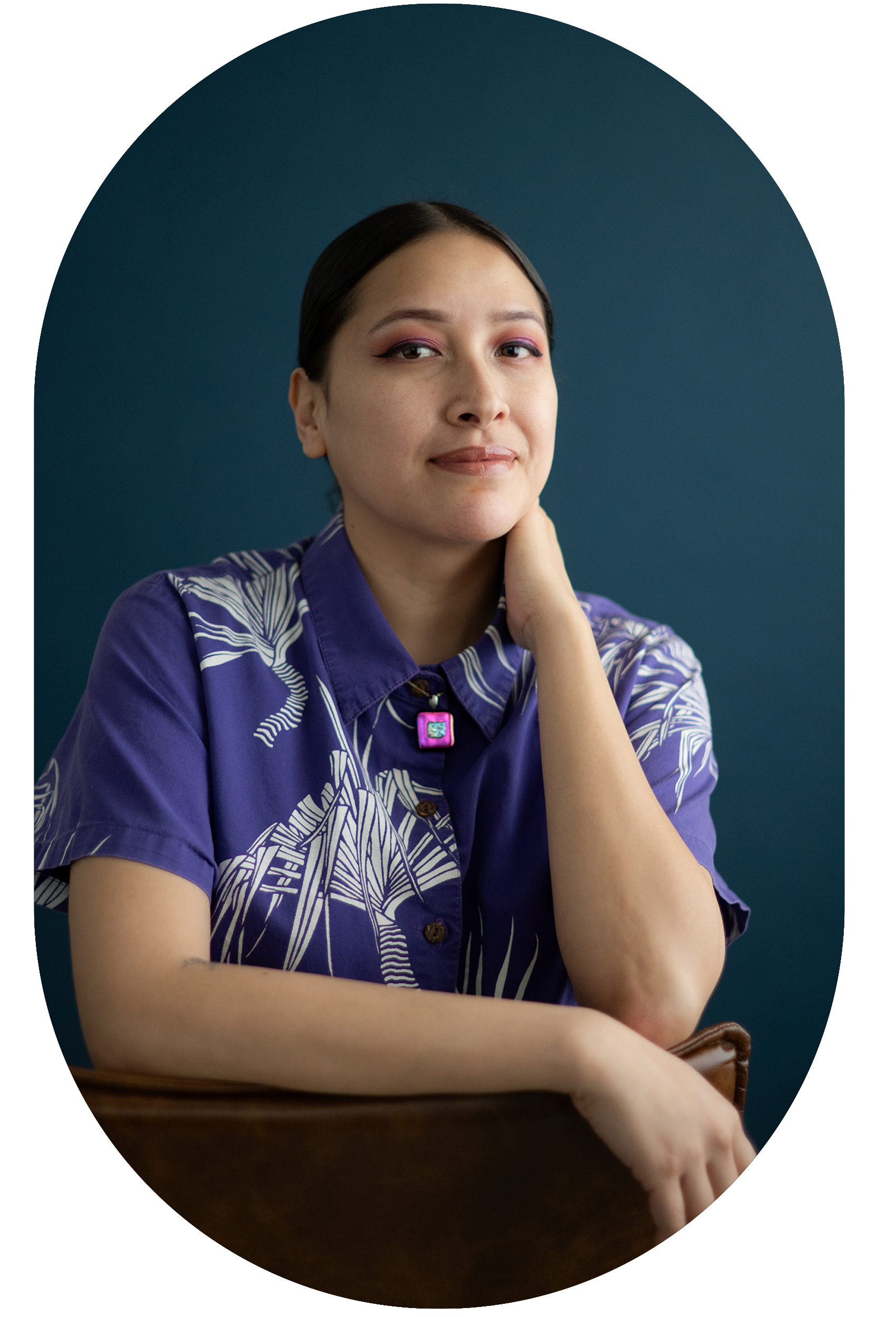 A person with dark hair, wearing pink eyeshadow and a purple shirt with a leaf pattern, sitting against a dark teal background with her hand resting on her neck, looking at the camera.