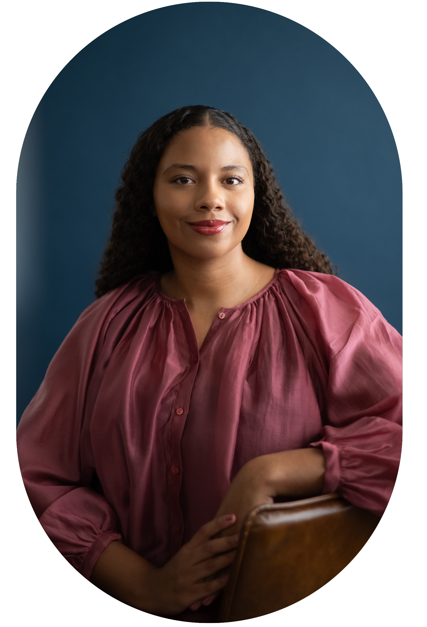 A person with curly hair wearing a pink satin blouse, smiling, seated against a blue background.