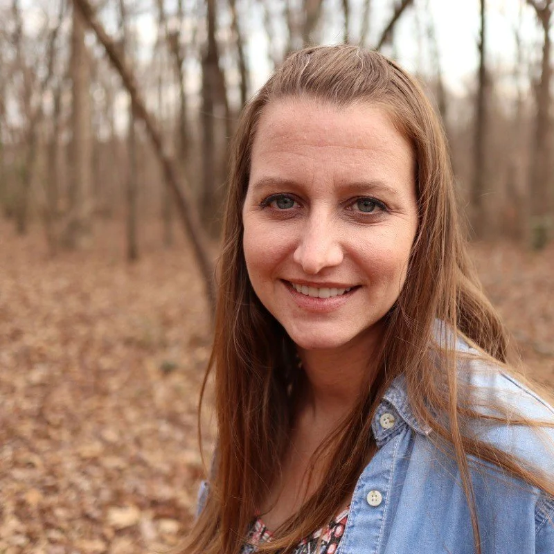 A woman with long brown hair smiling outdoors in a wooded area with fallen leaves on the ground.