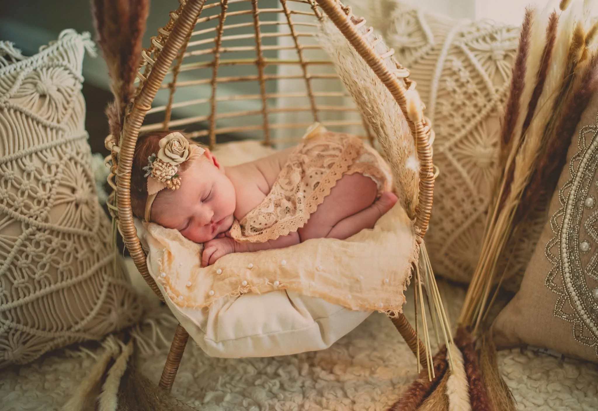 A sleeping baby girl with a floral headband, lying in a wicker cradle surrounded by cream-colored crochet pillows and decorative dried flowers.