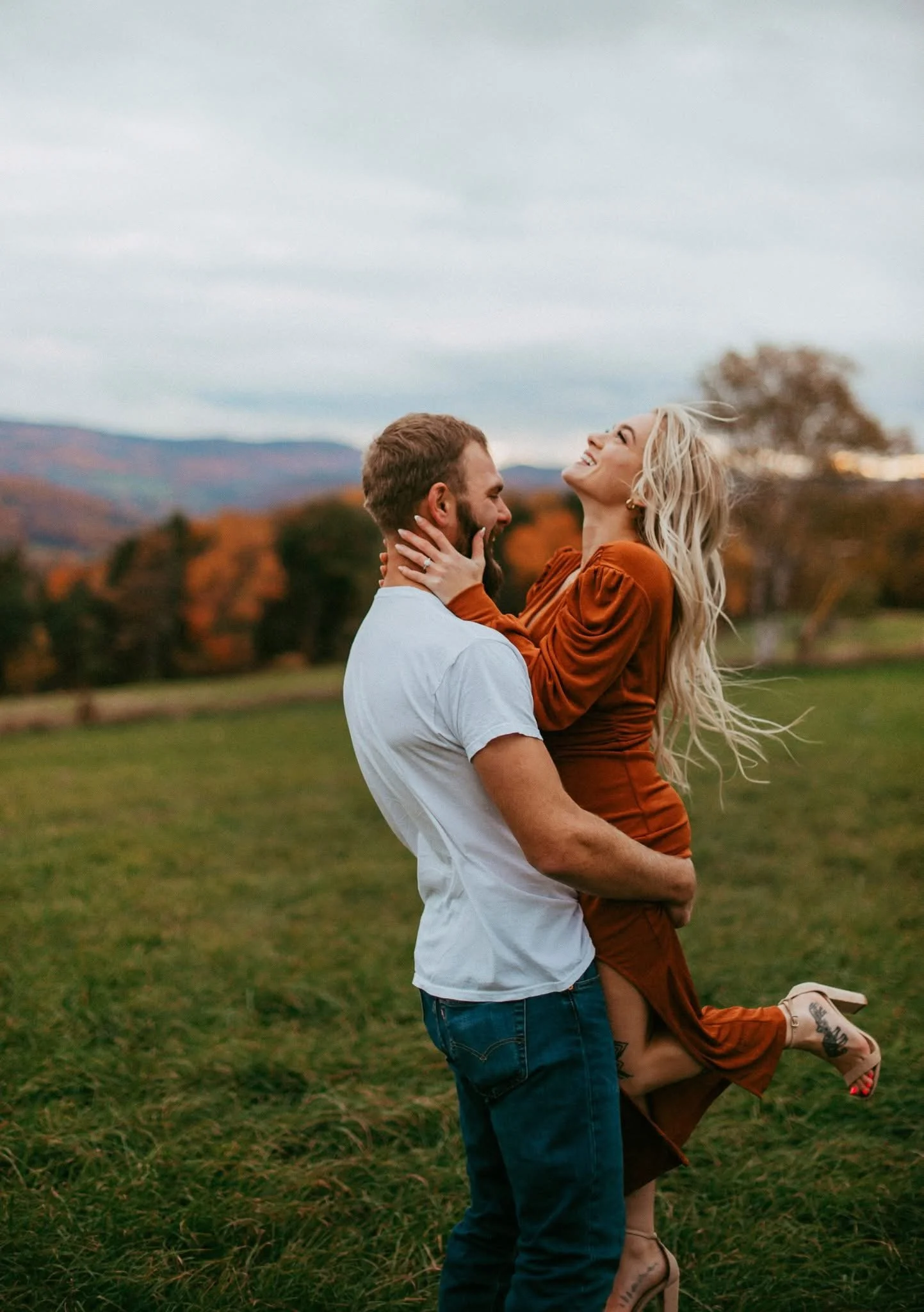 A man holding a woman in a field during autumn, both smiling and laughing. The woman has long blonde hair, is wearing an orange dress and high heels. The man is wearing a white t-shirt and jeans.