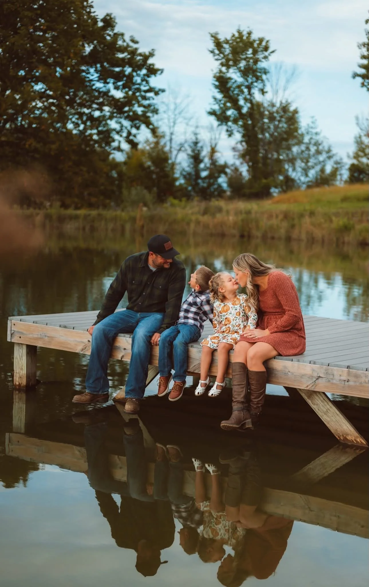 A family of four sitting on a wooden dock by a body of water, with trees and a cloudy sky in the background. The family includes two adults and two children, with the children receiving a kiss from the woman.