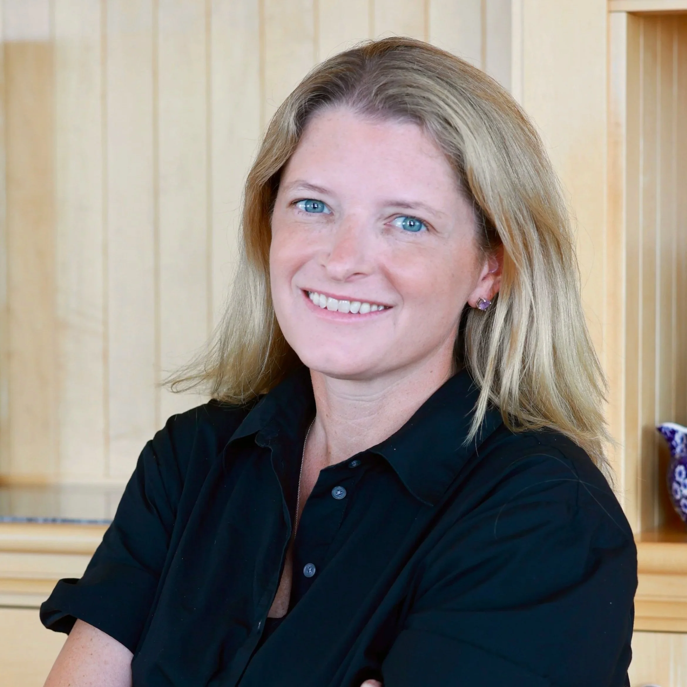 A woman with long blonde hair and blue eyes smiling, wearing a black collared shirt, in front of a wooden background.
