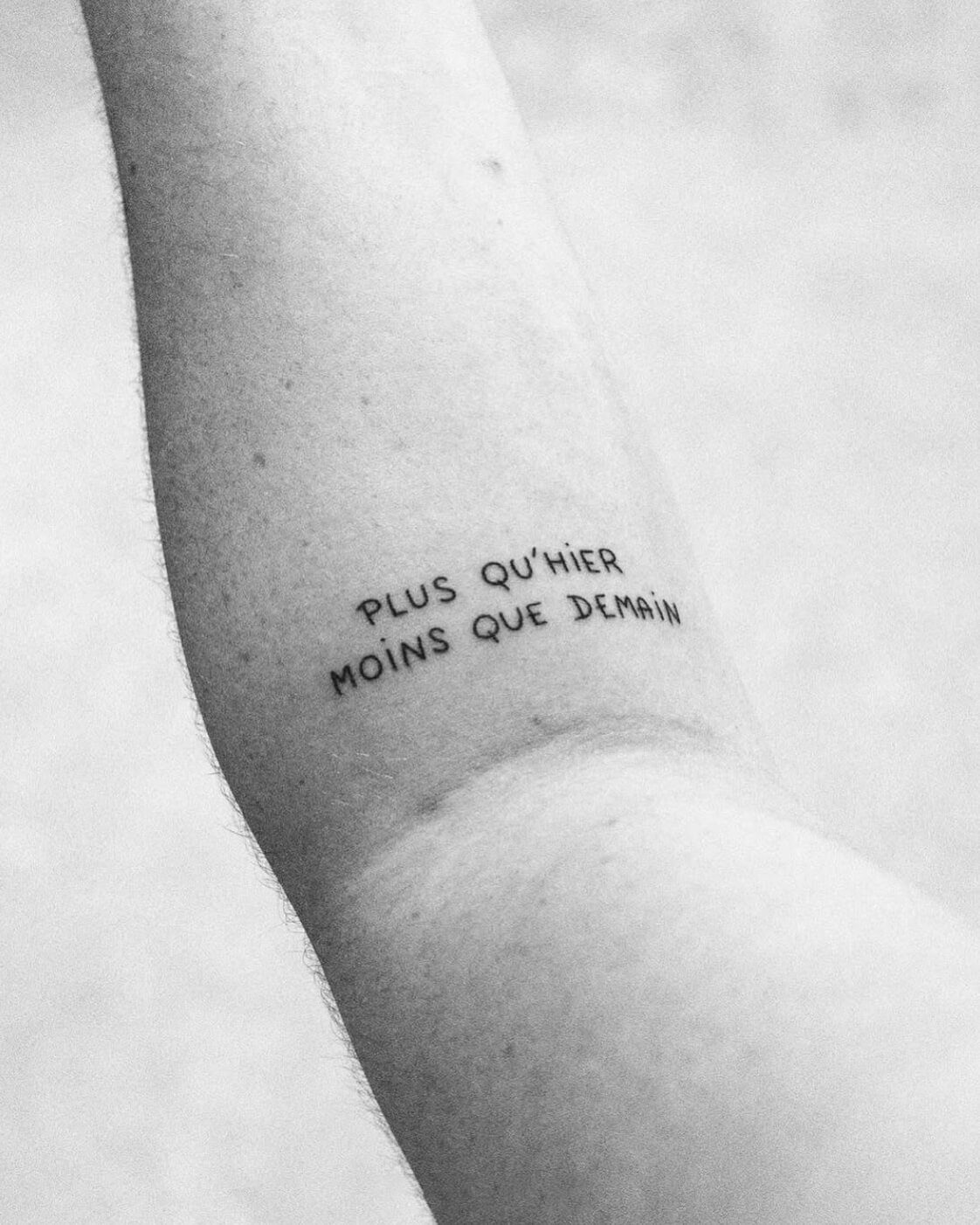 Tatouage sur un bras avec l'inscription en français "Plus qu'hier, moins que demain".