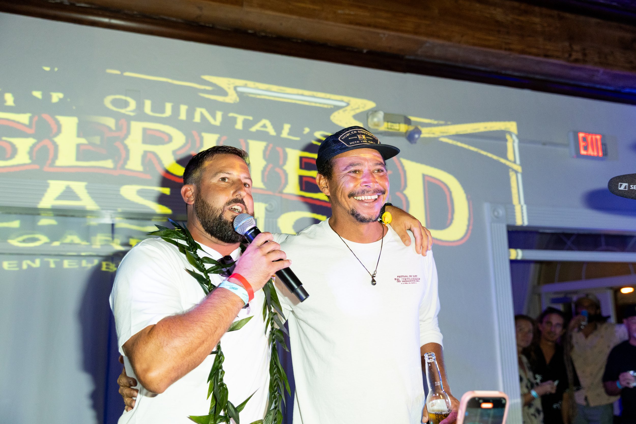 Two men stand close together, smiling; the man on the left is speaking into a microphone and is wearing a white t-shirt and a lei, while the man on the right is wearing a baseball cap and a white shirt. Behind them is a projection or sign with bright