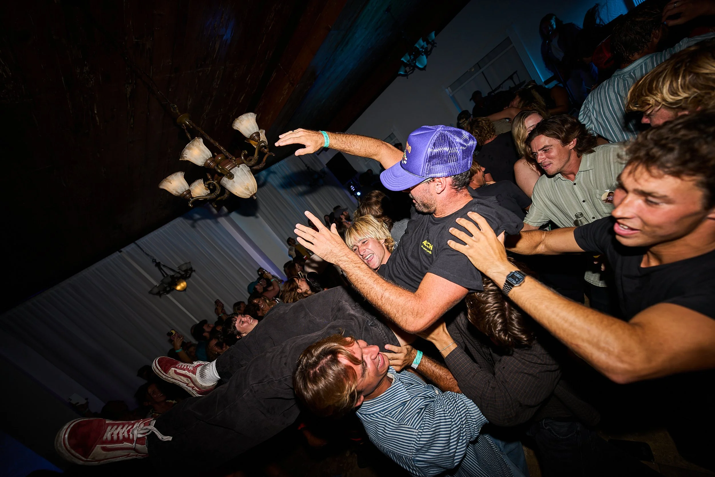 People at a party or concert standing close together, some with arms around each other, and dancing or enjoying the event, with a dark indoor setting and a chandelier hanging from the ceiling.