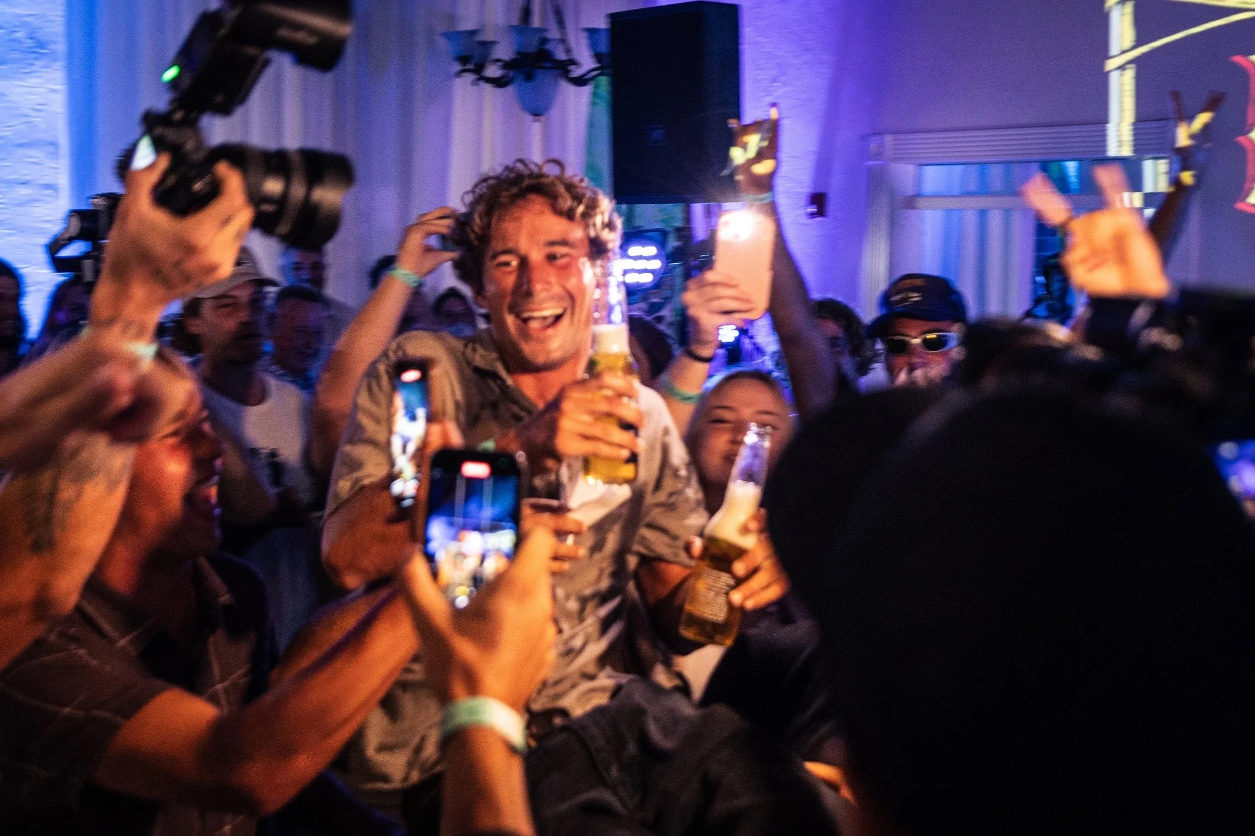 A group of people at a lively event or party, with one person in the center holding a beer and smiling, surrounded by others taking photos and enjoying the moment. The background features colorful lighting and decorations.
