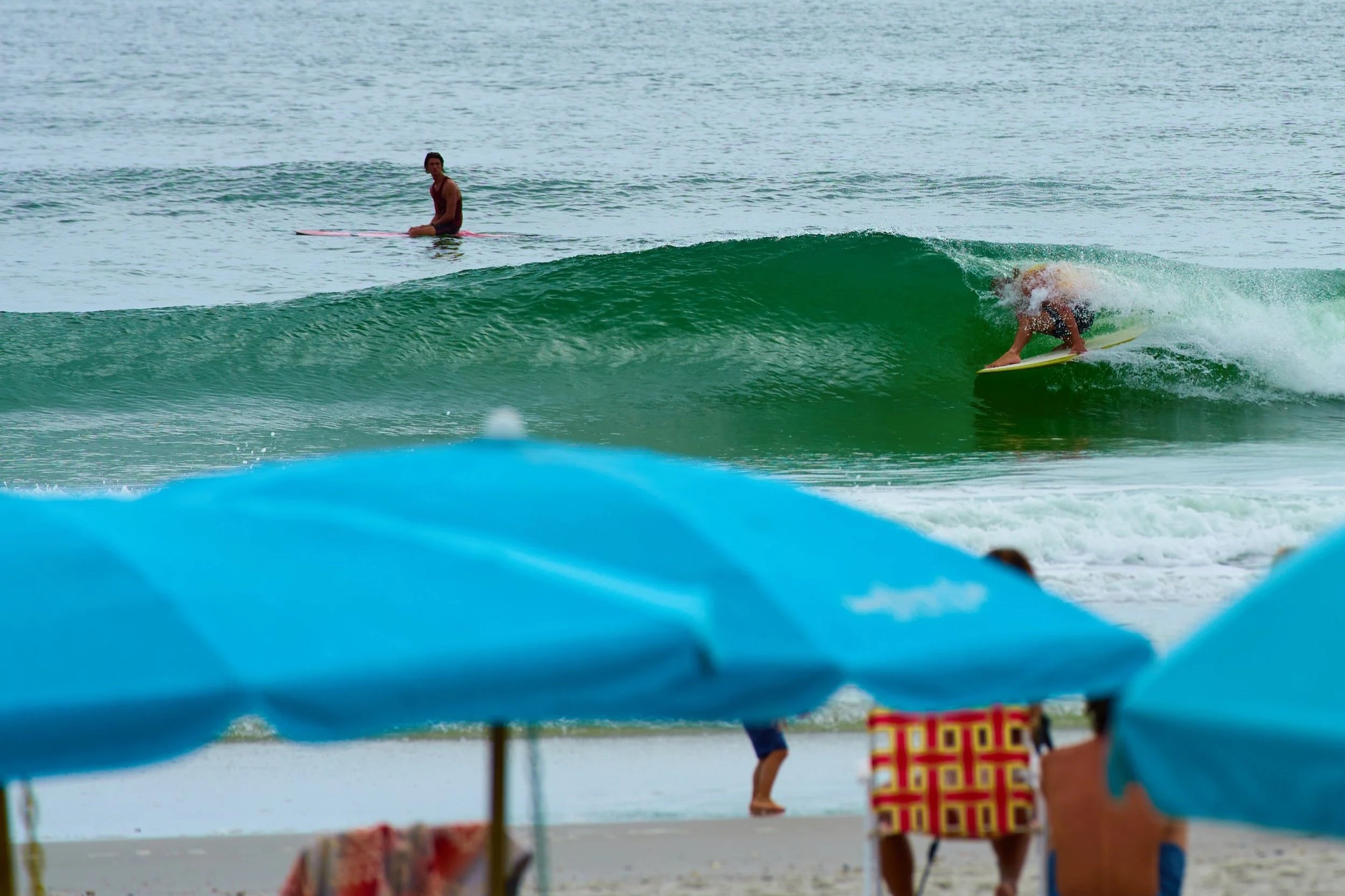 A person surfing on a wave at the beach with another person sitting on a surfboard in the water nearby, and beach umbrellas and people on the sand in the background.