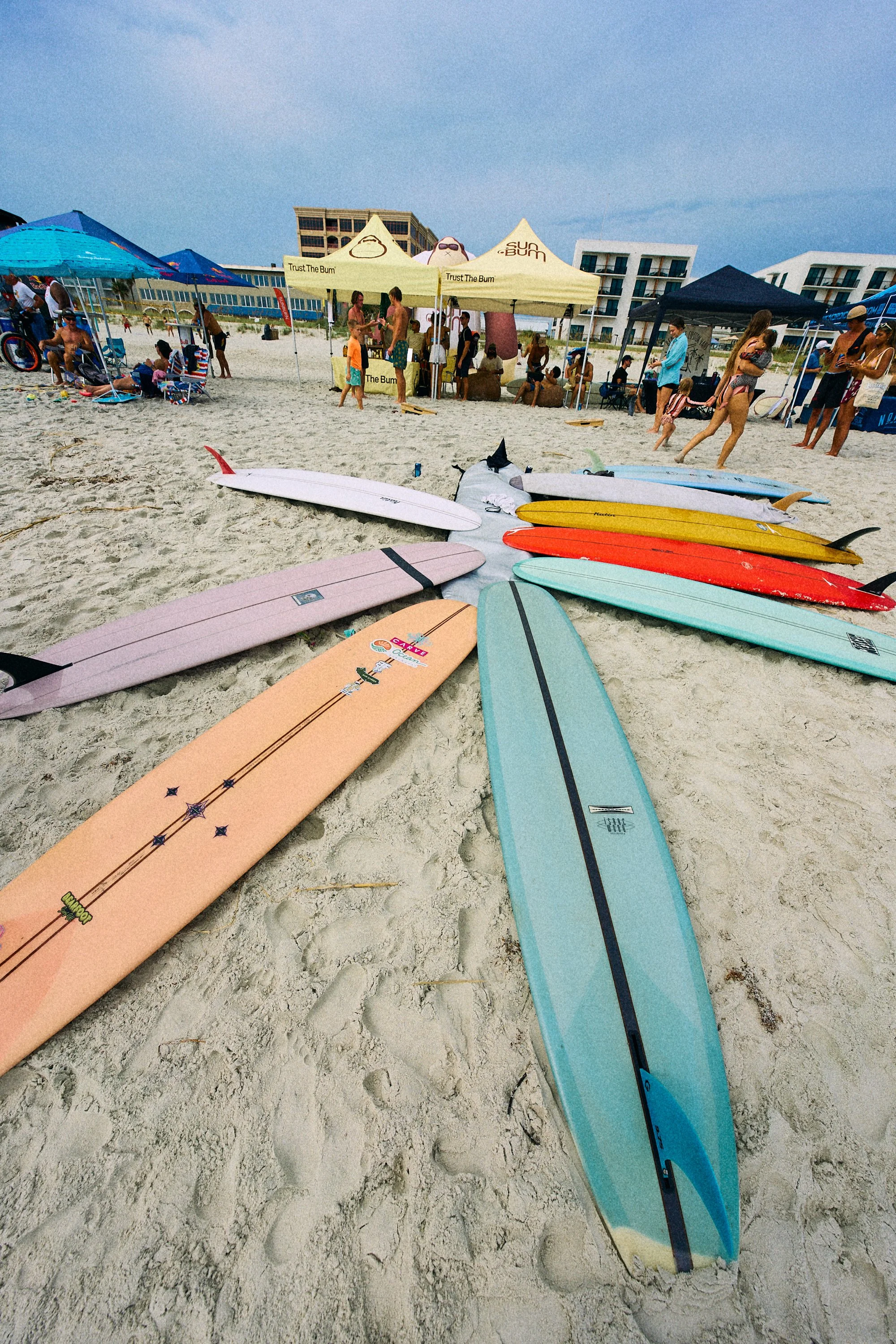 Multiple colorful surfboards on the sandy beach with tents and people in the background.