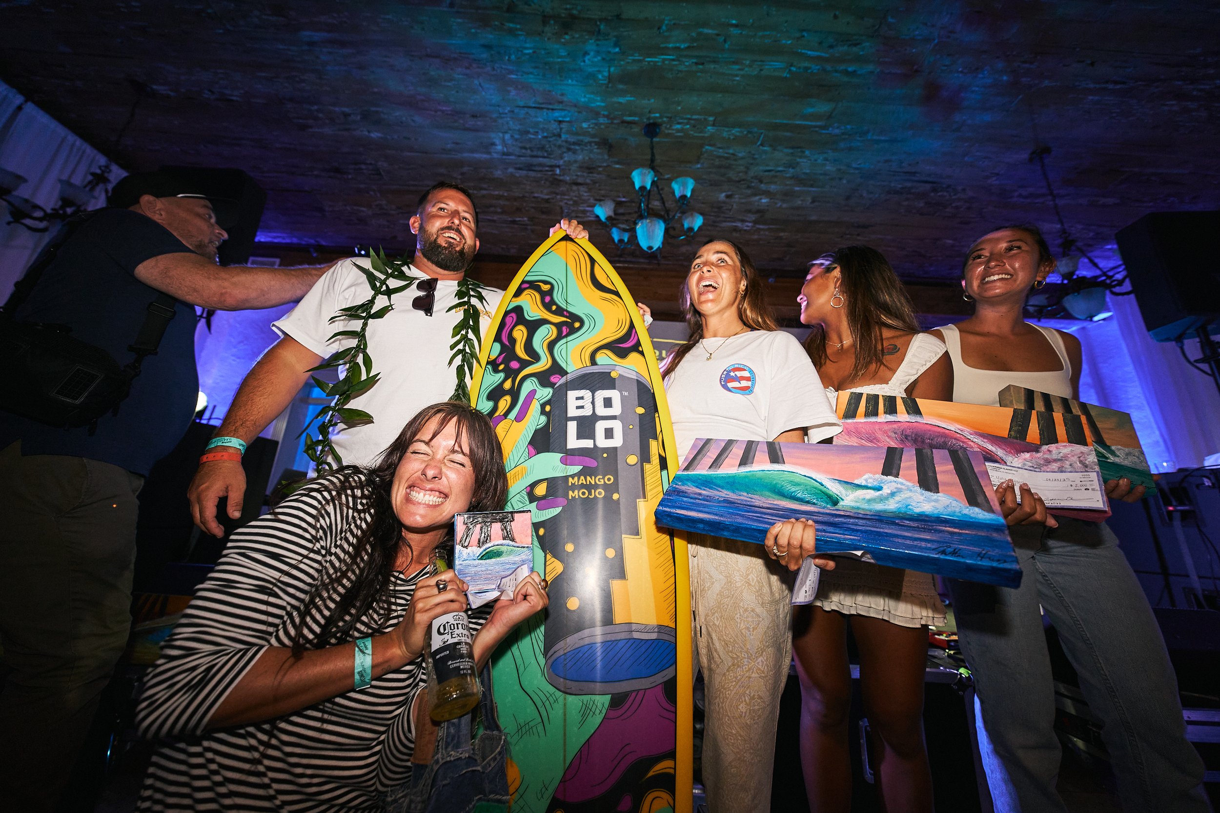 Group of people celebrating indoors, some holding art paintings, with a surfboard display in the center, colorful lighting, and smiling faces.