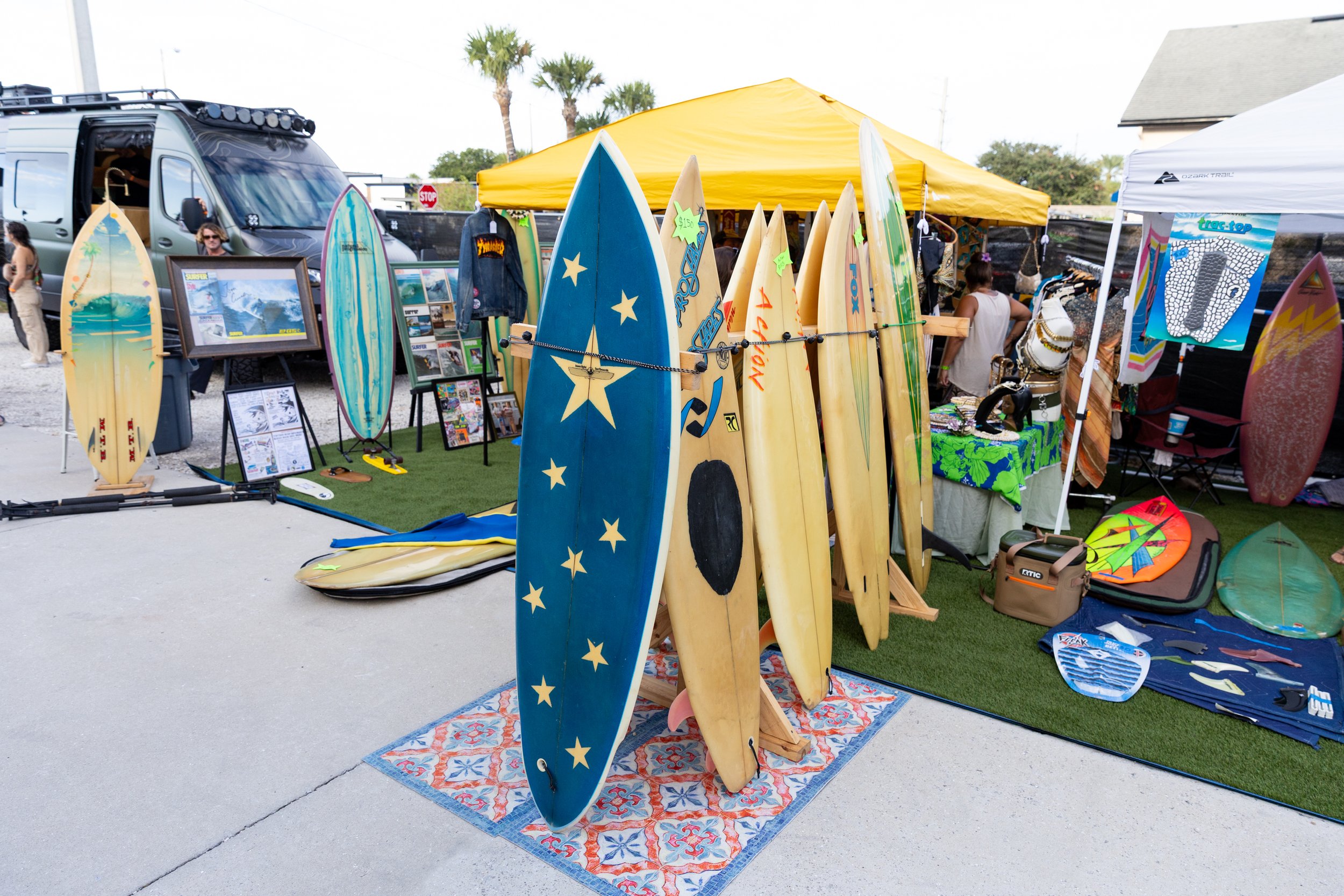 Outdoor market stall displaying various surfboards, some with colorful designs, standing on a decorative rug. Behind the surfboards are framed pictures, clothing, and other items for sale under a yellow canopy tent. People are browsing the items, wit