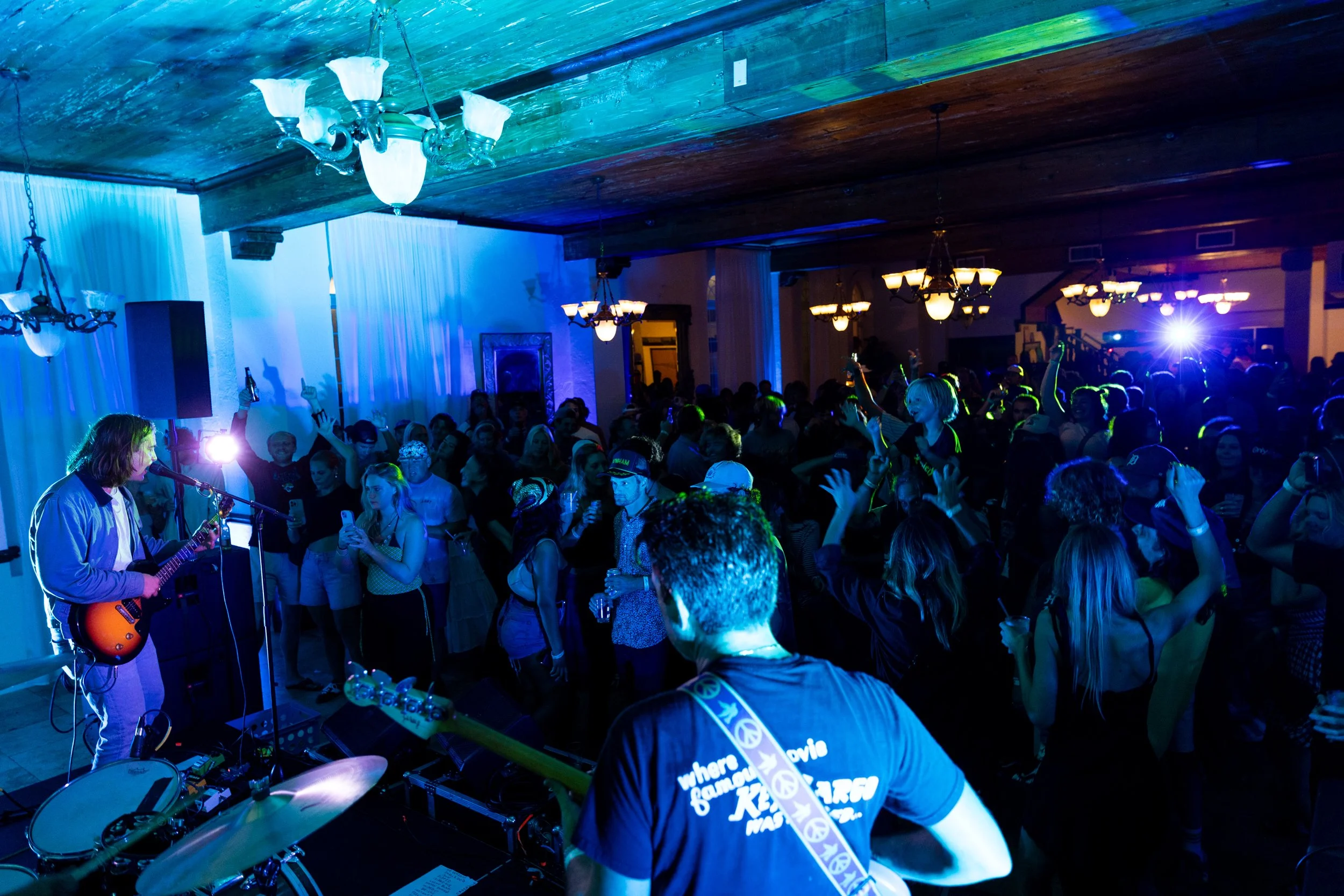 A live music event with a guitarist performing on stage in front of a large crowd, colorful lighting, chandeliers, and people dancing and singing along.