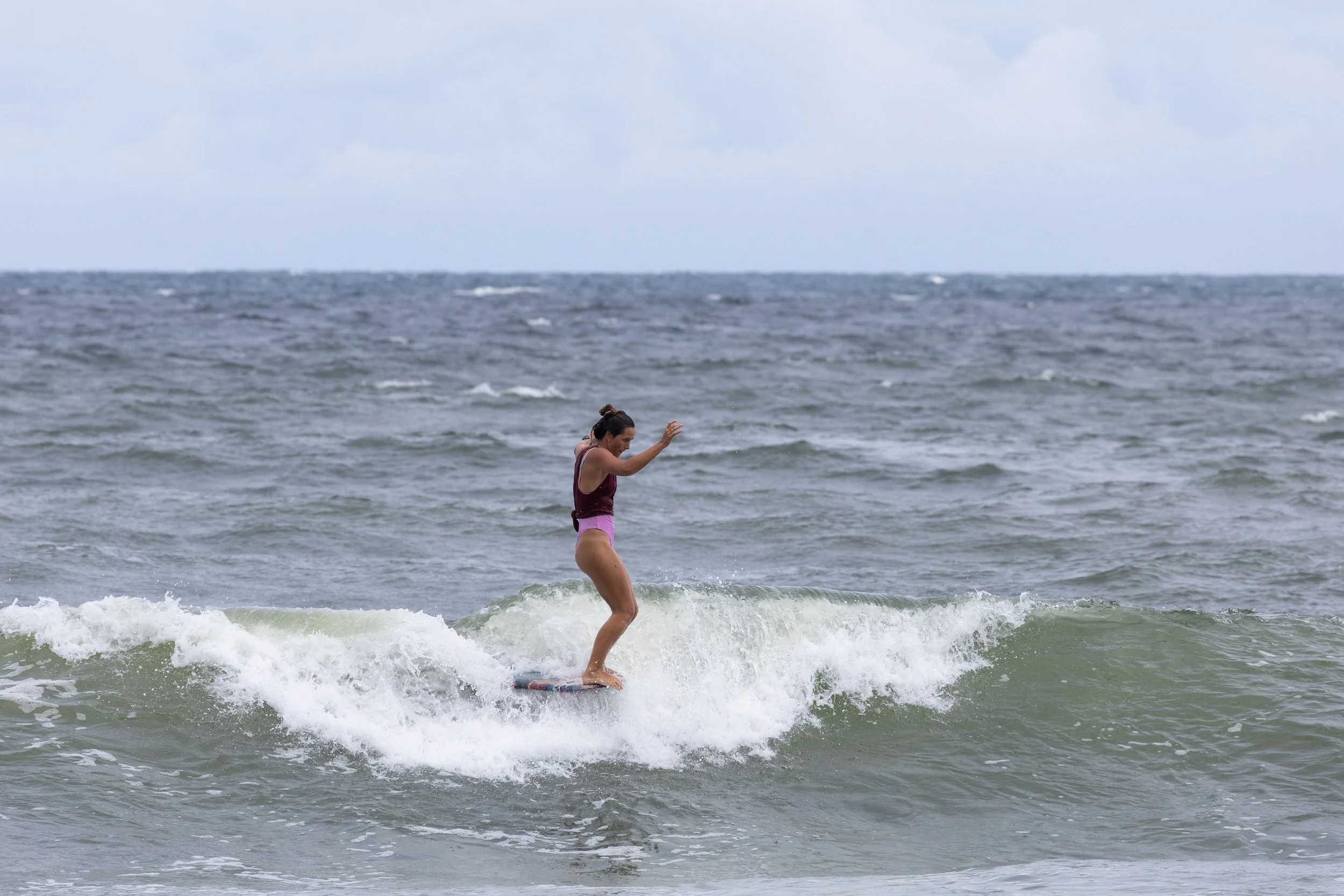A woman in a maroon tank top and pink shorts balancing on a surfboard riding a small wave in the ocean under a cloudy sky.