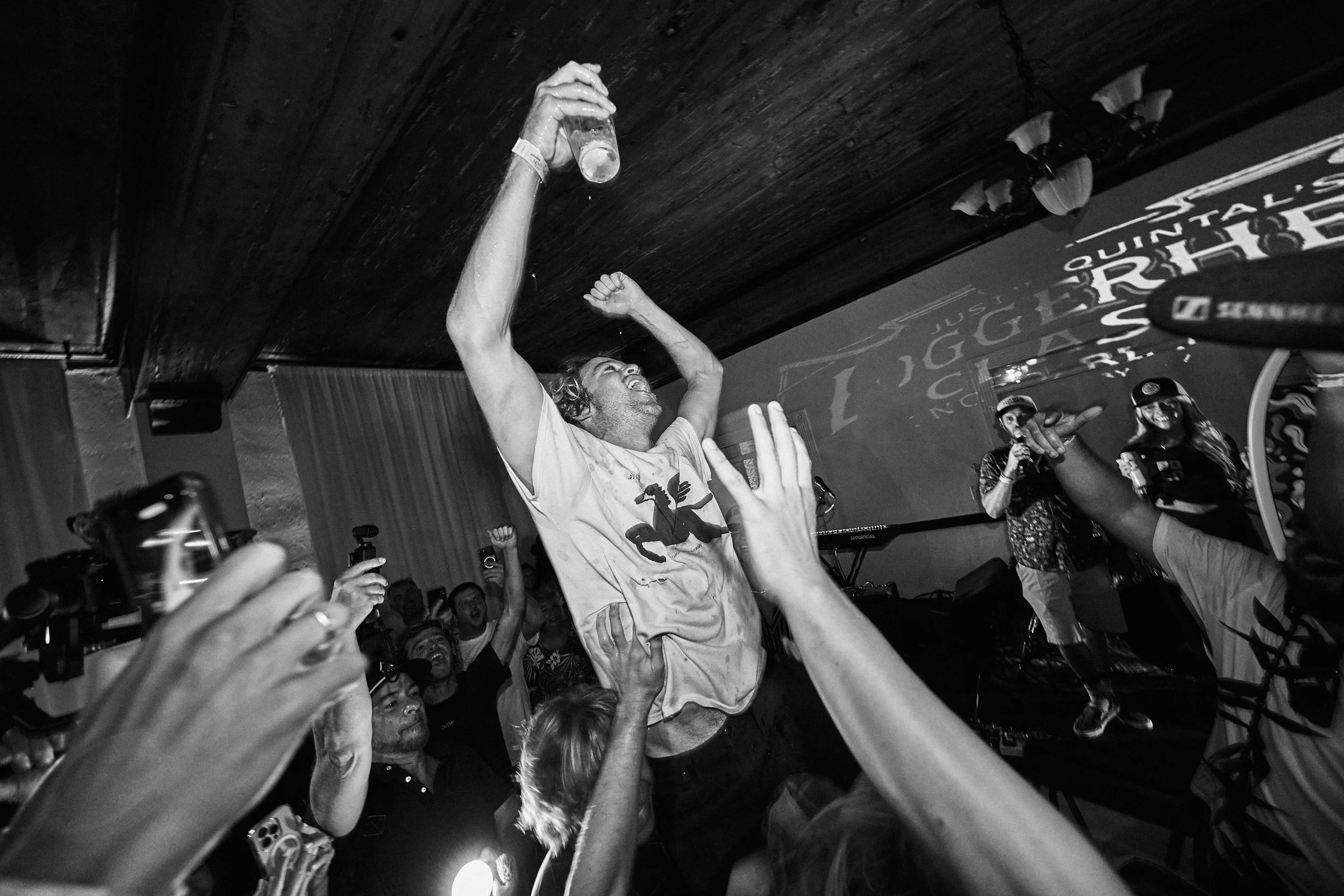 A man is raising a glass in celebration while crowd surfing at a lively party or concert. People around him are cheering and reaching out, with hands and phones visible. The scene is indoors with a wooden ceiling and some wall decor.