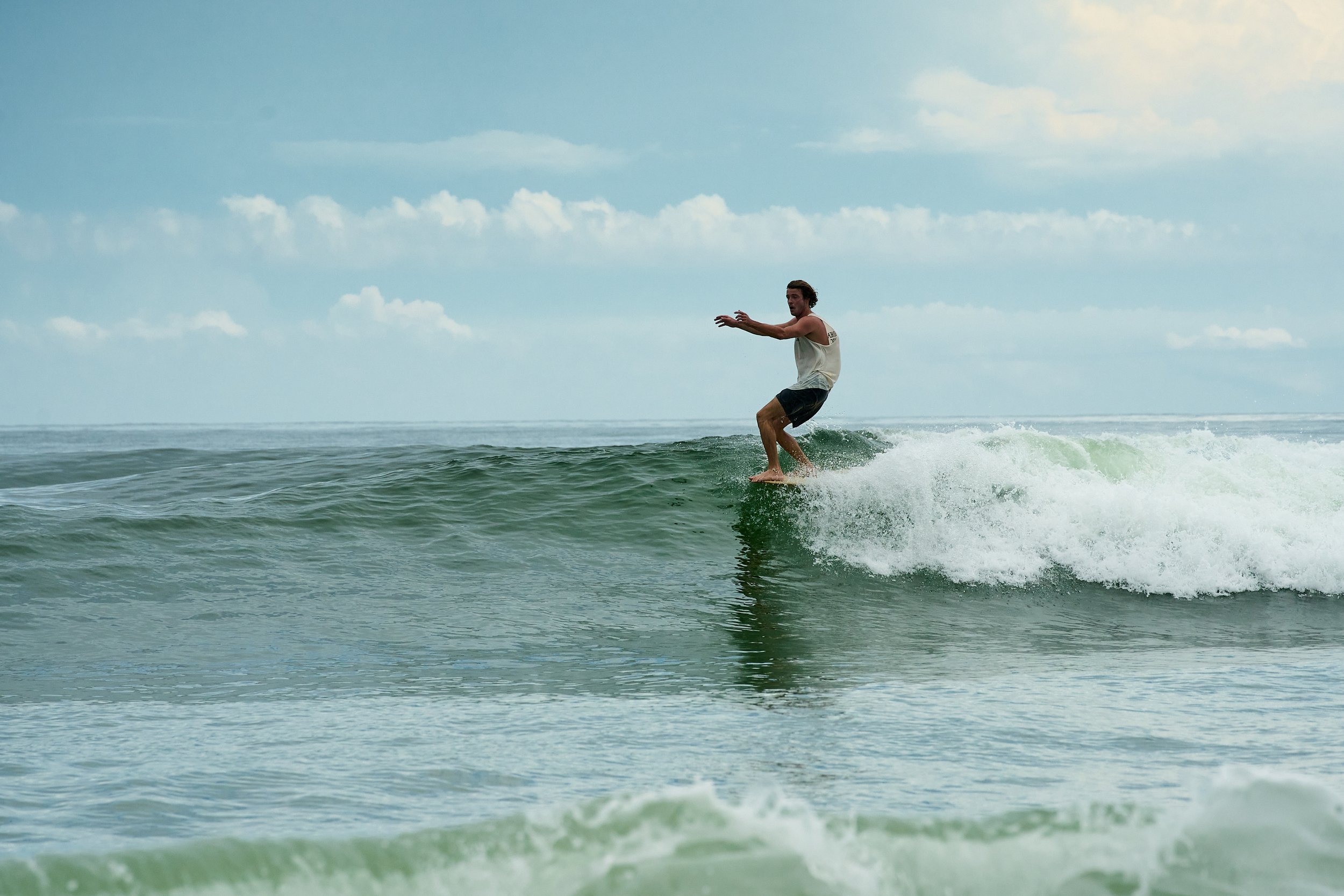 A man surfing on a small wave in the ocean, wearing a sleeveless shirt and shorts, with a cloudy sky overhead.