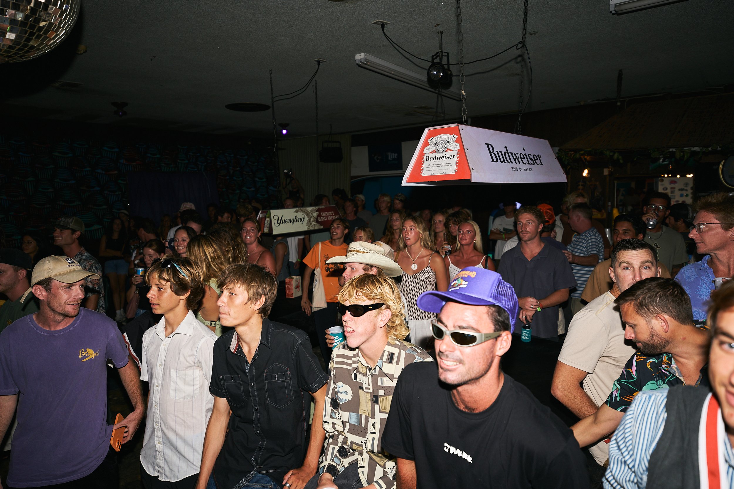 Crowd of people in a dimly lit venue, some holding drinks, with a prominent Budweiser sign hanging from the ceiling.