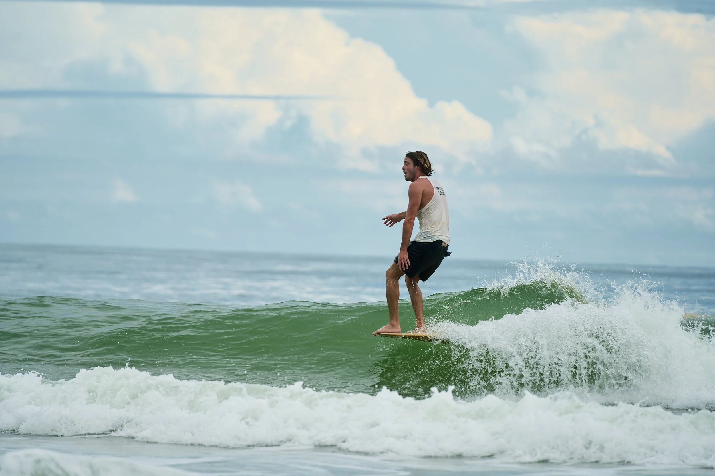 A man with long hair surfing on a wave in the ocean under a partly cloudy sky.