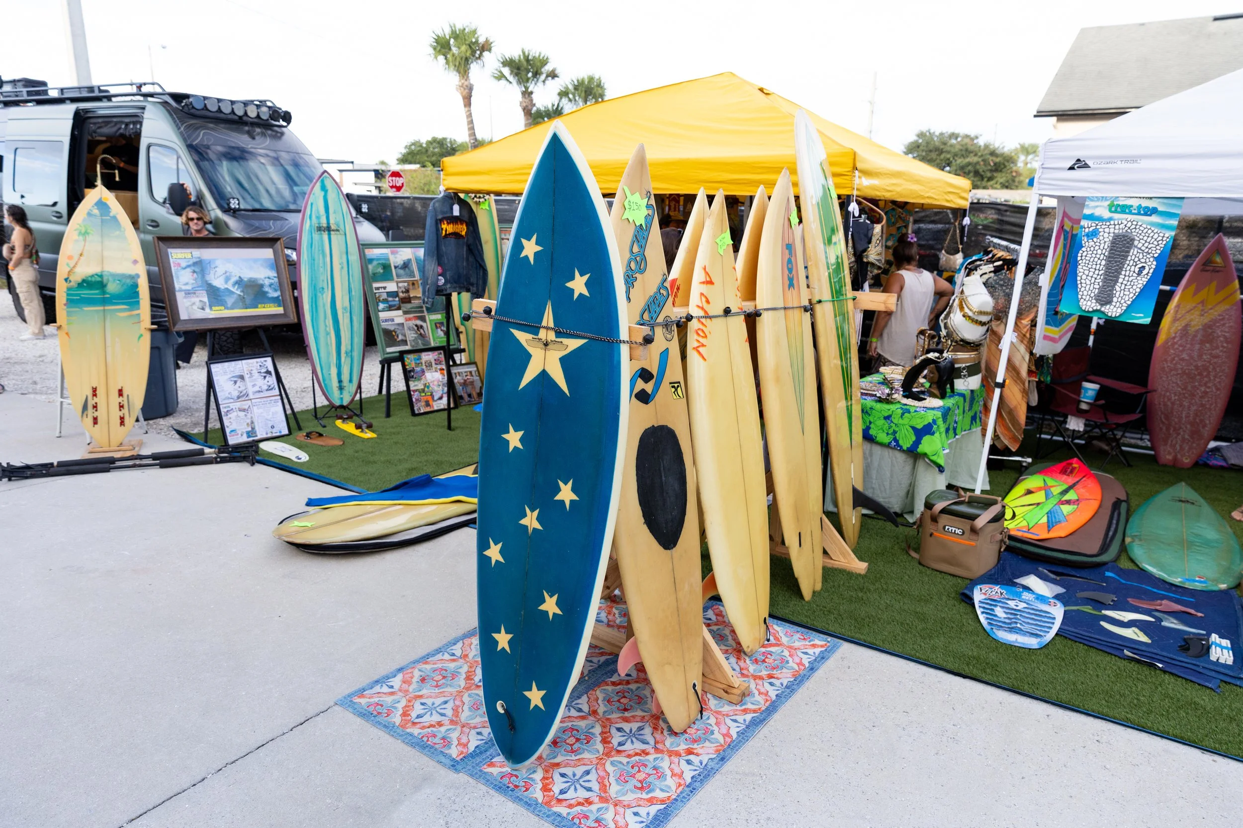 Outdoor market stall selling surfboards, with a yellow canopy, palm trees in the background, and various items including framed pictures, clothing, and beach accessories.
