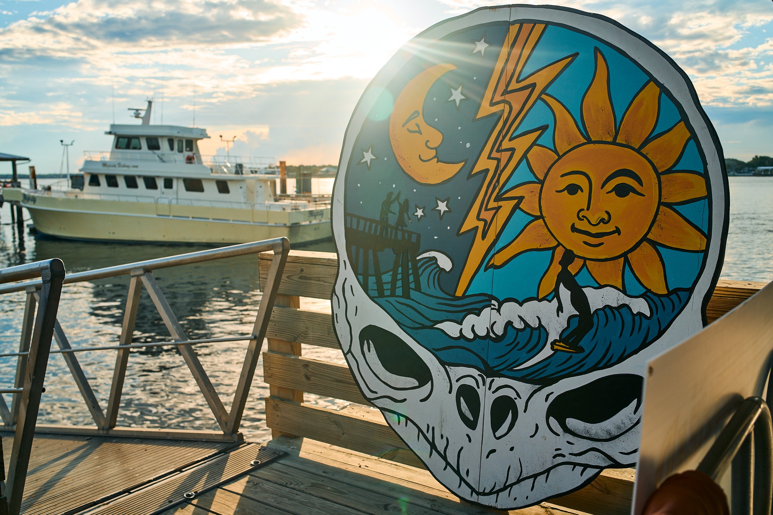 A colorful sign featuring a sun with a smiling face, a moon with a face, stars, and lightning bolts, situated near a dock with a boat in the background during sunset.