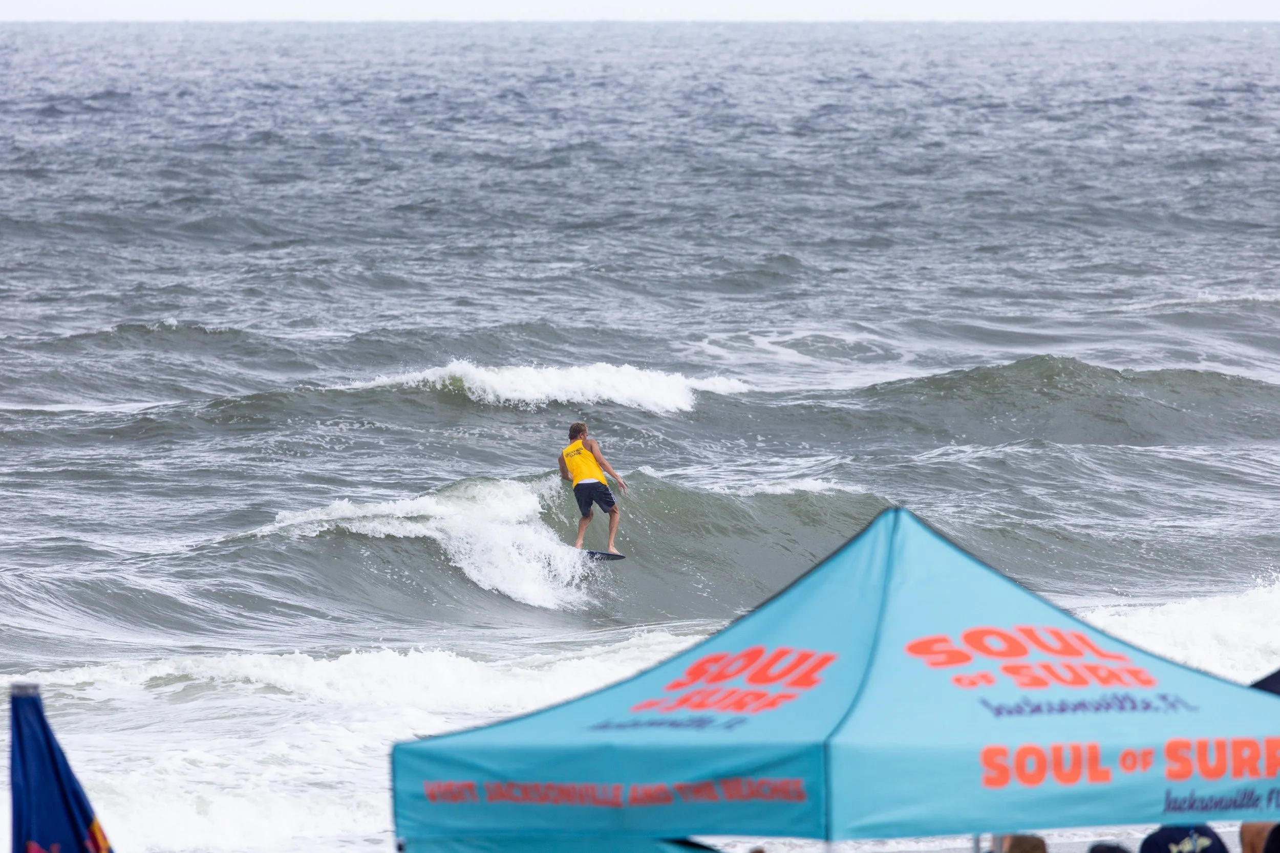 Surfer riding a wave in the ocean, seen from under a blue umbrella with orange and black text, overcast sky in the background.