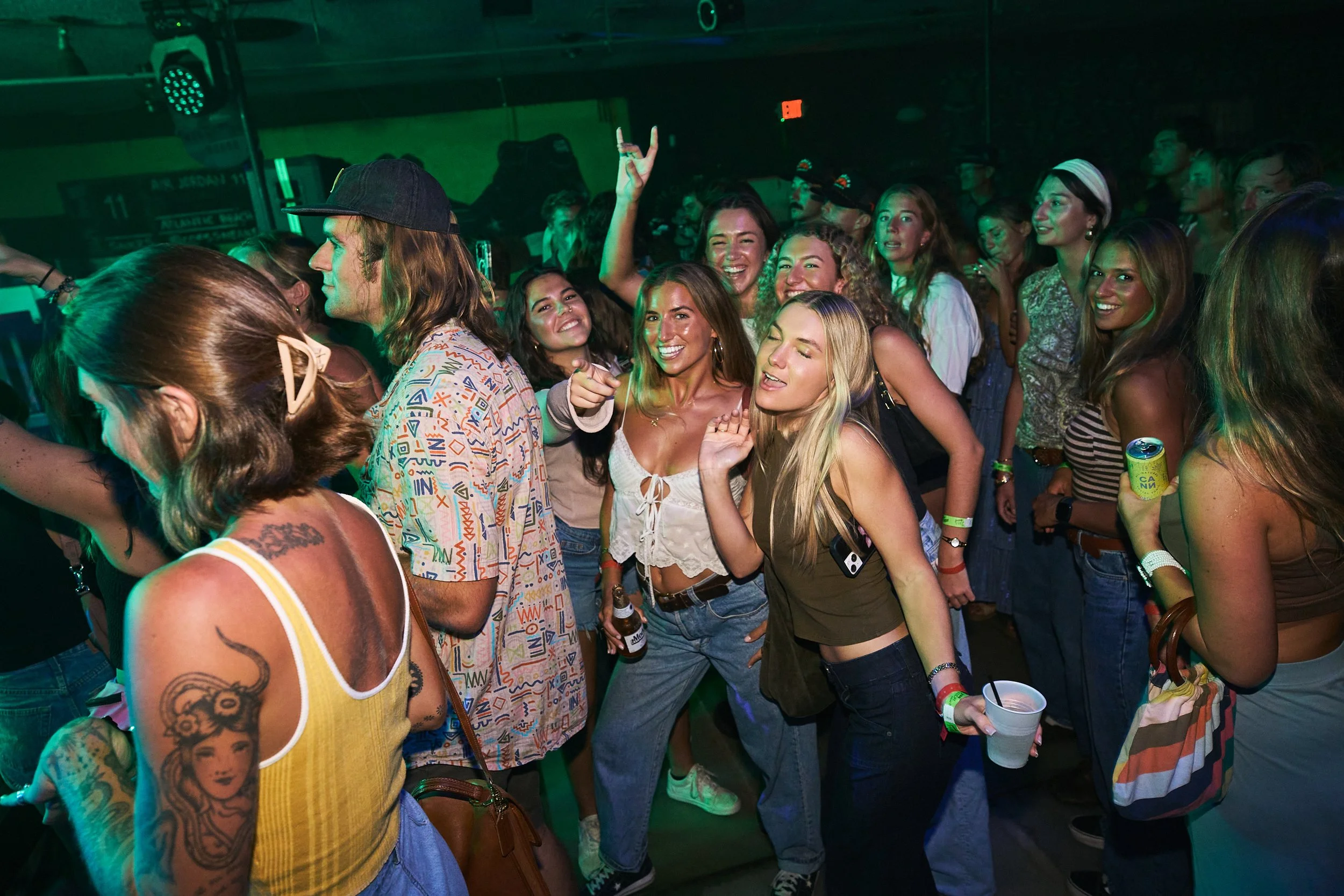 People at a lively indoor party or concert, smiling, dancing, and holding drinks, with colorful lighting and a dark background.