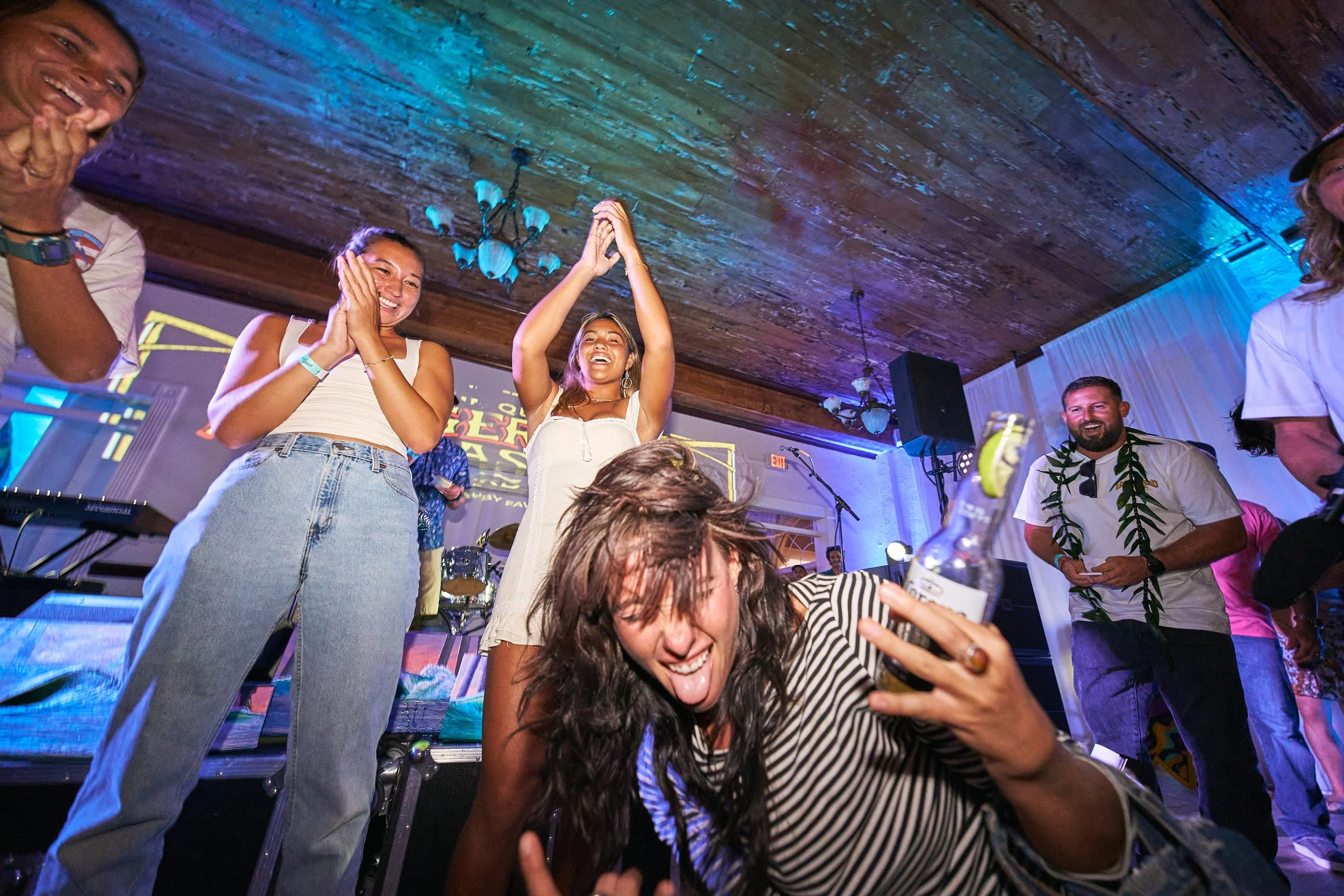 People at a lively party or concert, smiling and enjoying music. Two women are standing, clapping and smiling, while a person in a striped shirt is sitting in front, sticking out their tongue. The ceiling has wooden planks with colorful lighting.
