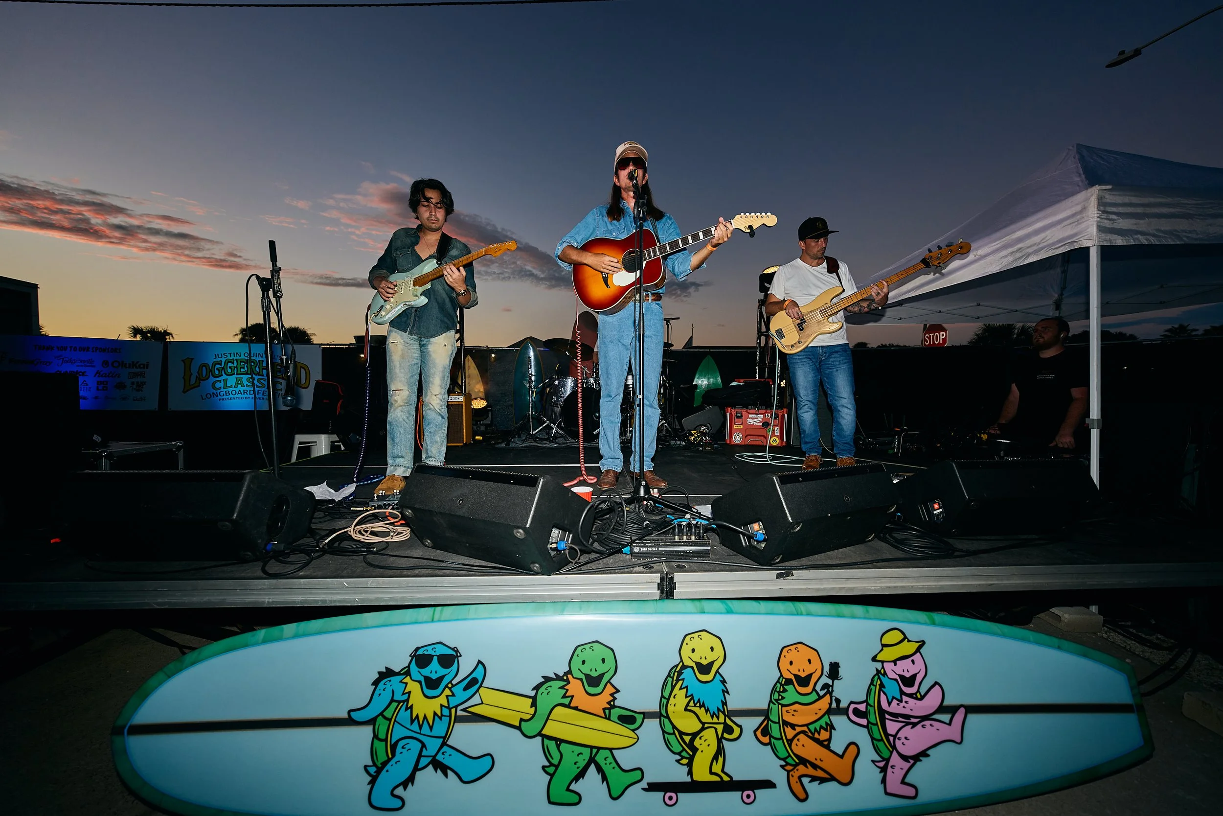 A band playing live on an outdoor stage at sunset, with three musicians holding guitars and singing into microphones, and a colorful skateboard deck featuring cartoon turtles in front of the stage.