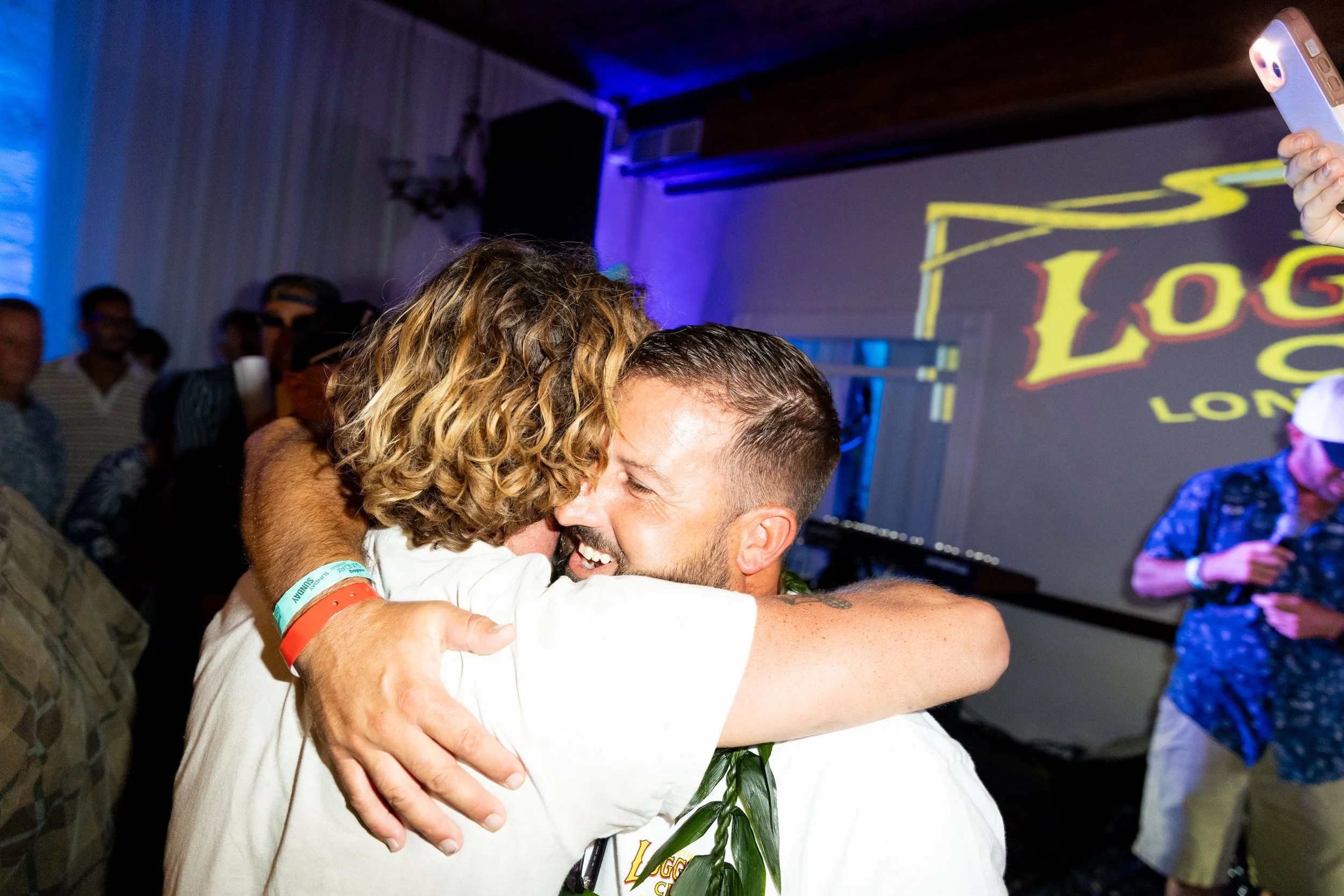 Two people hugging and smiling at a lively event, with multiple other attendees in the background and a large screen displaying colorful text on the wall.