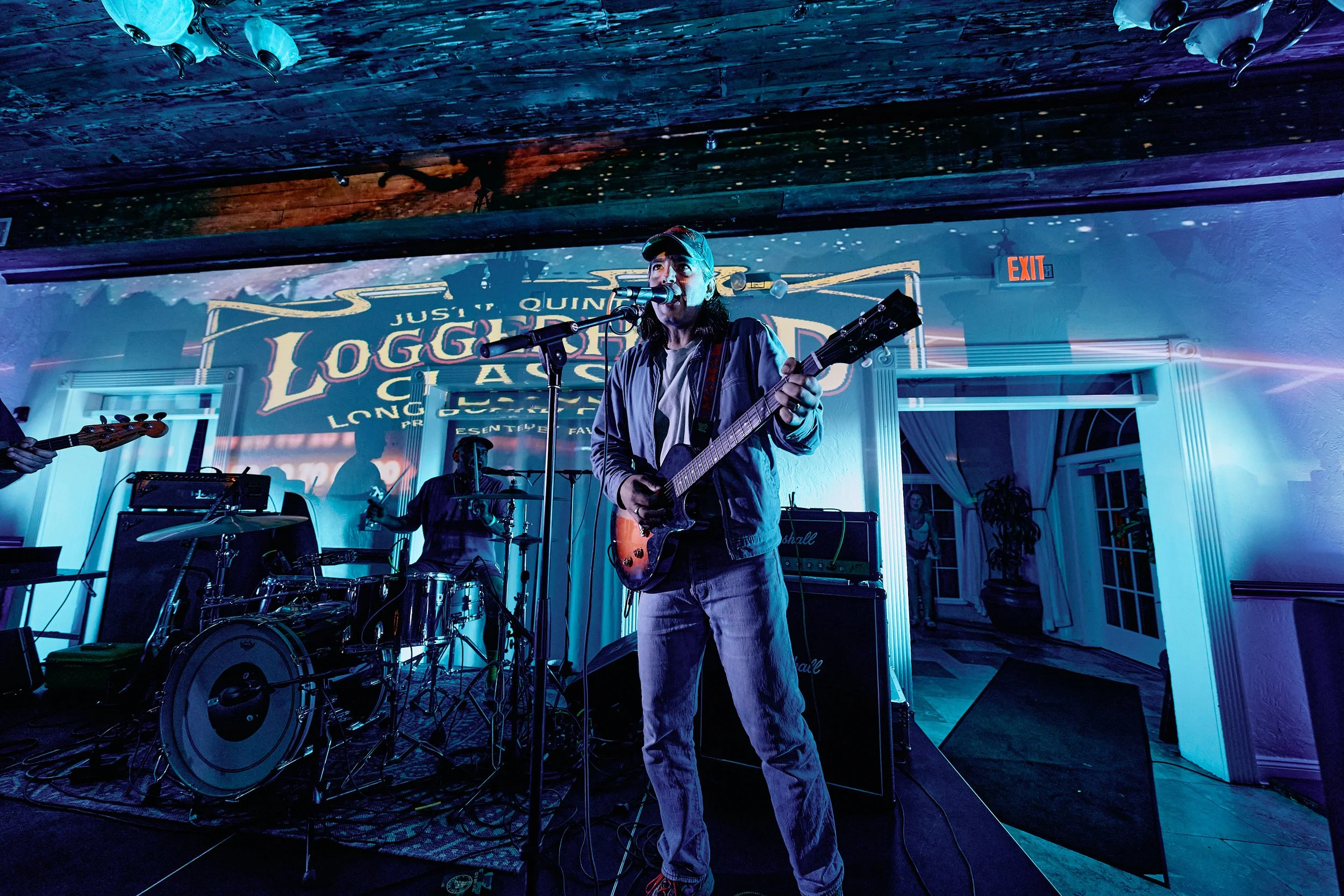 A musician on stage singing into a microphone while playing an electric guitar. The background features a lit sign and couple of windows with curtains. The scene is illuminated with blue and purple stage lights, and a drum set and other musical equip
