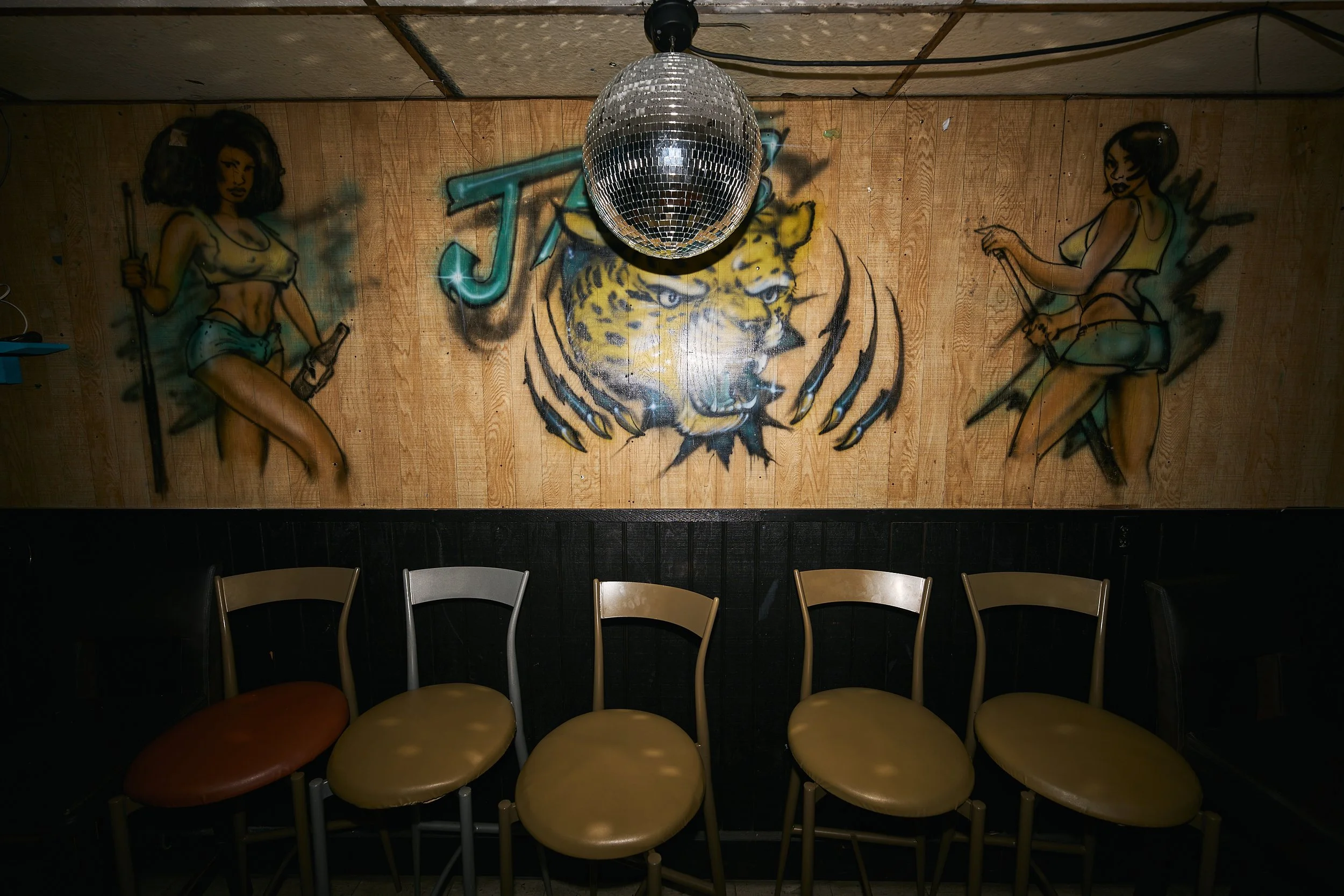 Graffiti mural on wooden wall featuring a central tiger face and two women with tattoos and provocative poses on either side, under a disco ball.