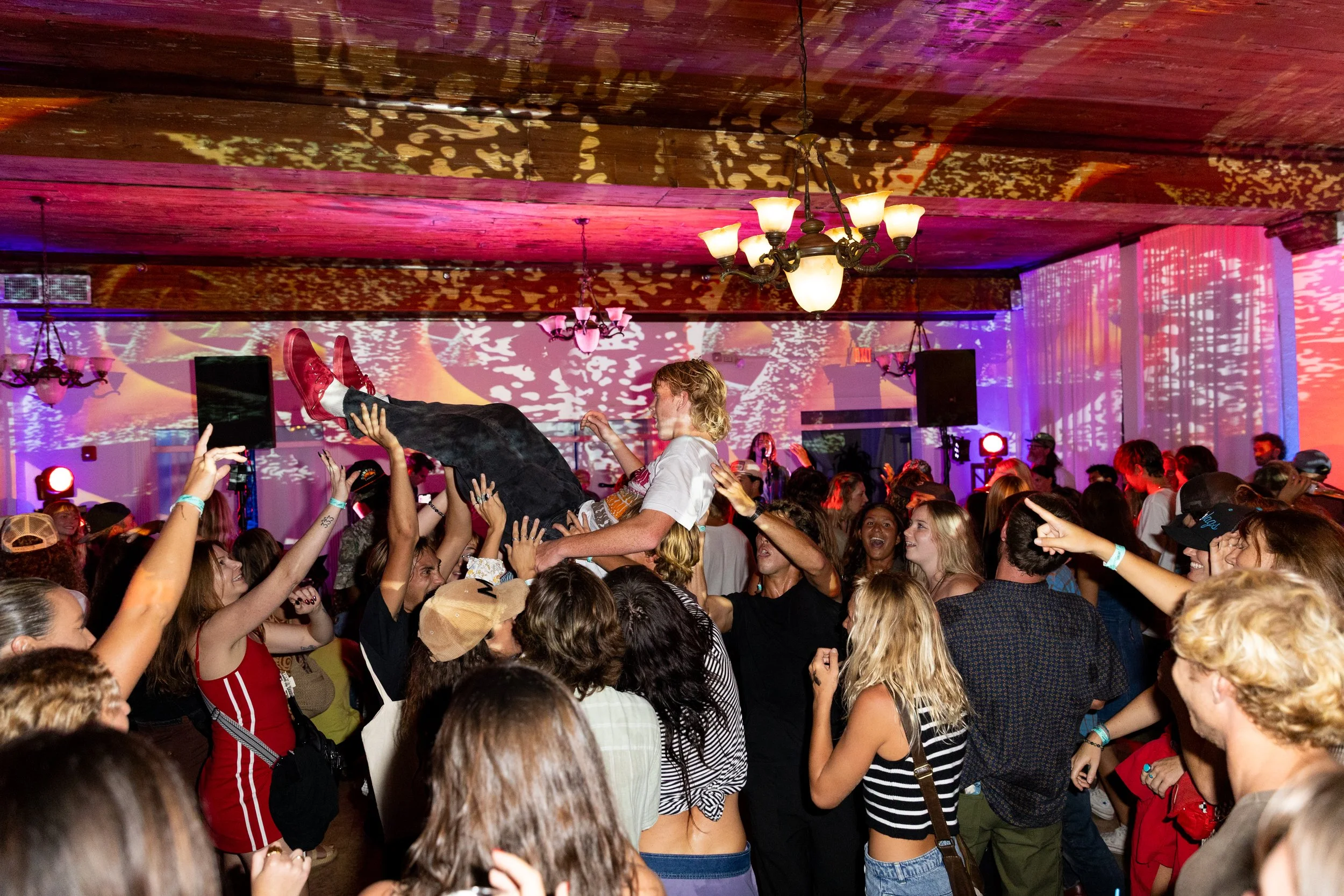 A crowd of people dancing and celebrating at an indoor event, with a person being lifted above the crowd. The room has wooden ceiling beams, chandeliers, and dynamic lighting effects.