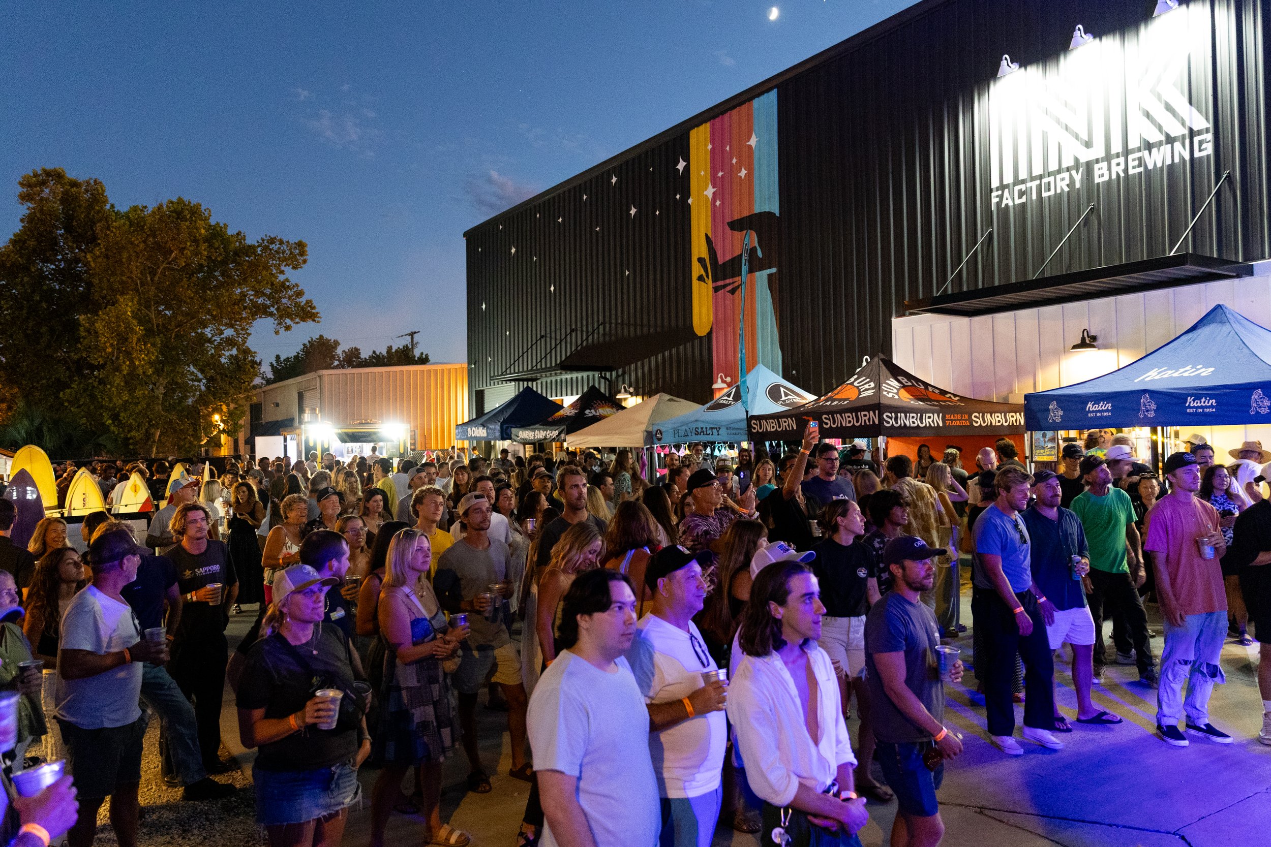 A large crowd of people gathered outdoors at night, attending an event in front of a building with a colorful mural and the logo of a brewery, under a clear sky with visible moon.