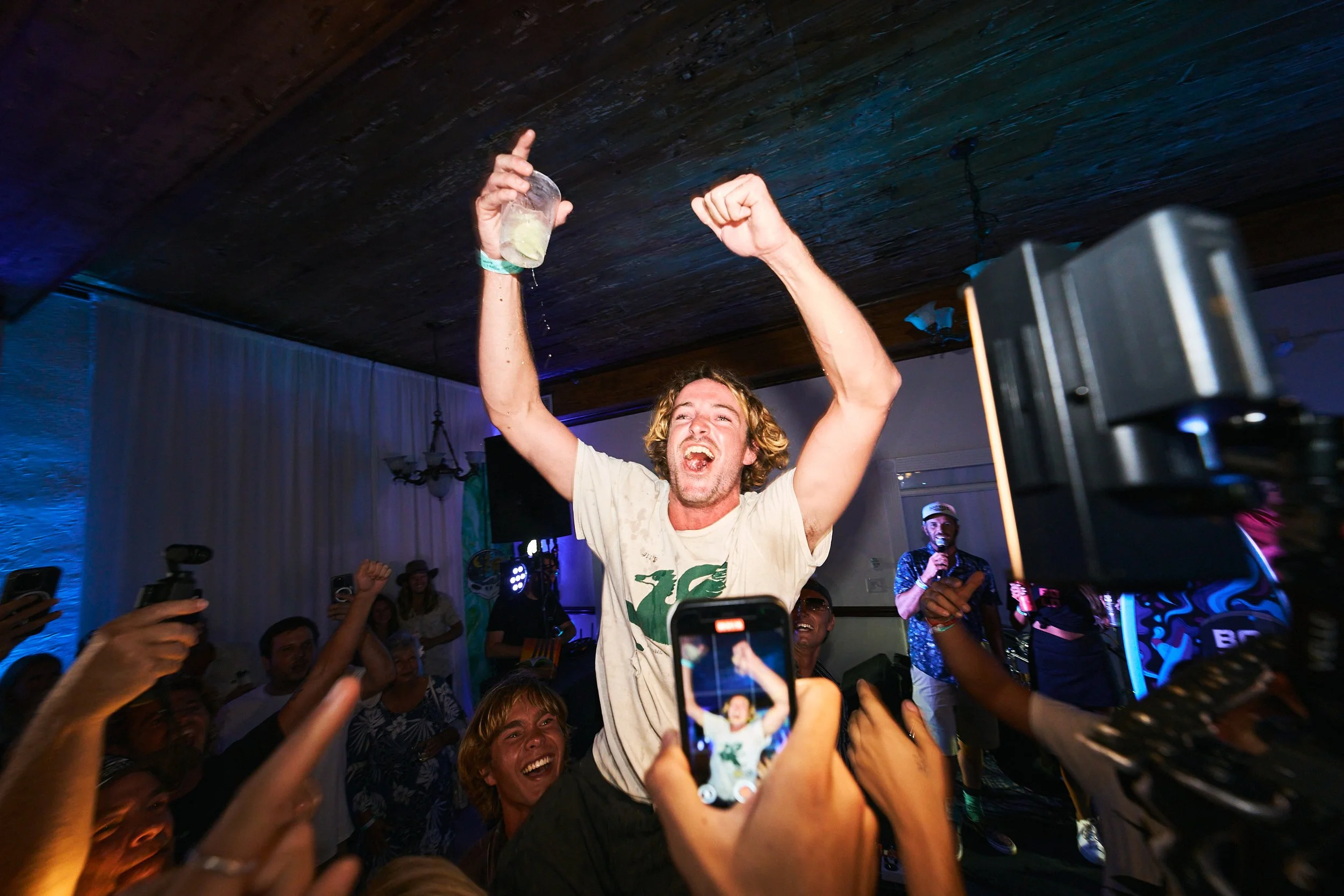 A man with curly blond hair and a white t-shirt is dancing and raising a plastic cup with liquid in it, surrounded by a crowd at a party. People are taking photos and videos with their phones. The setting appears to be a lively indoor event with colo
