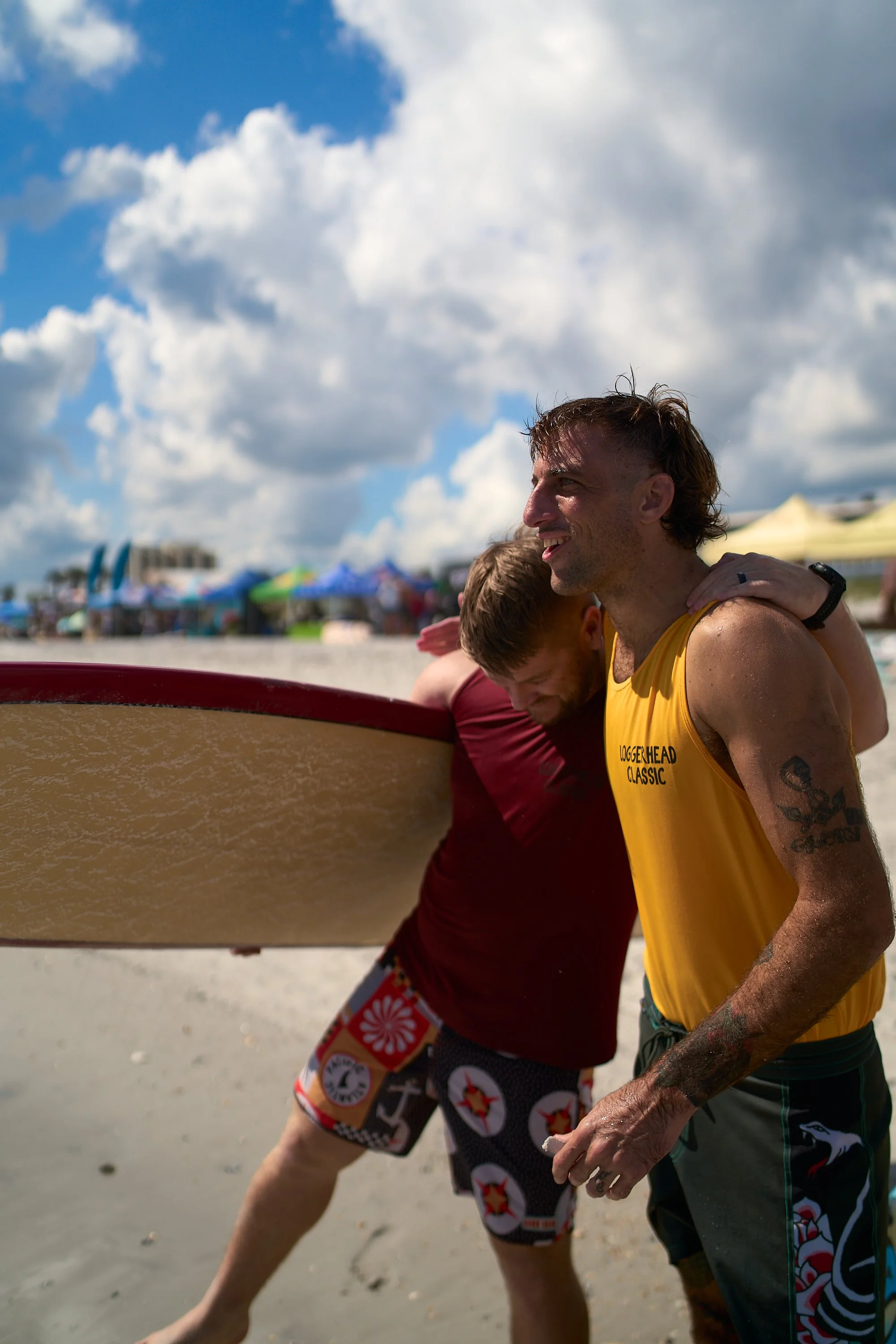 Two men at the beach holding a surfboard, with colorful umbrellas and tents in the background, under a partly cloudy sky.