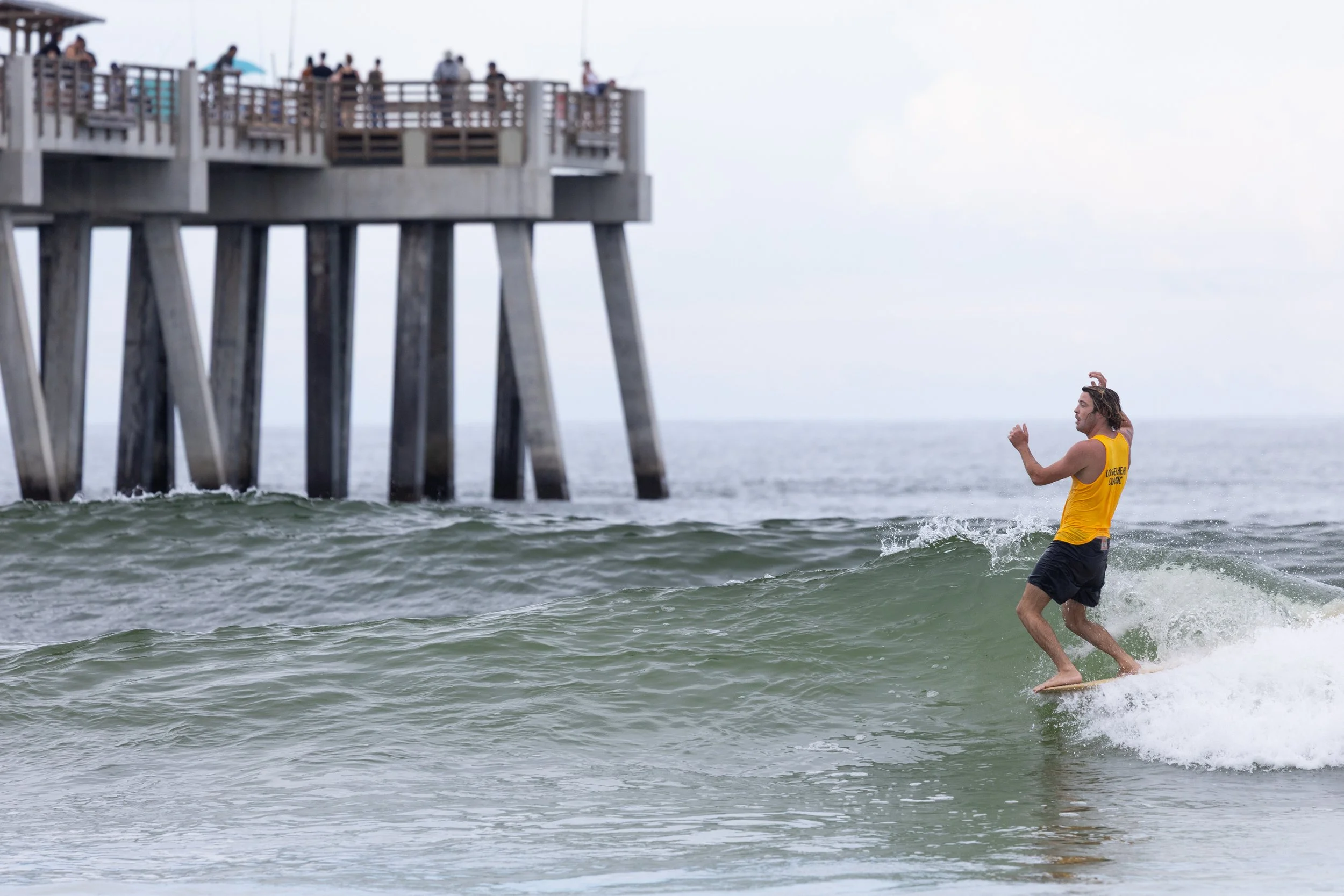 A man in a yellow tank top and black shorts is surfing in the ocean near a pier, with people on the pier observing.