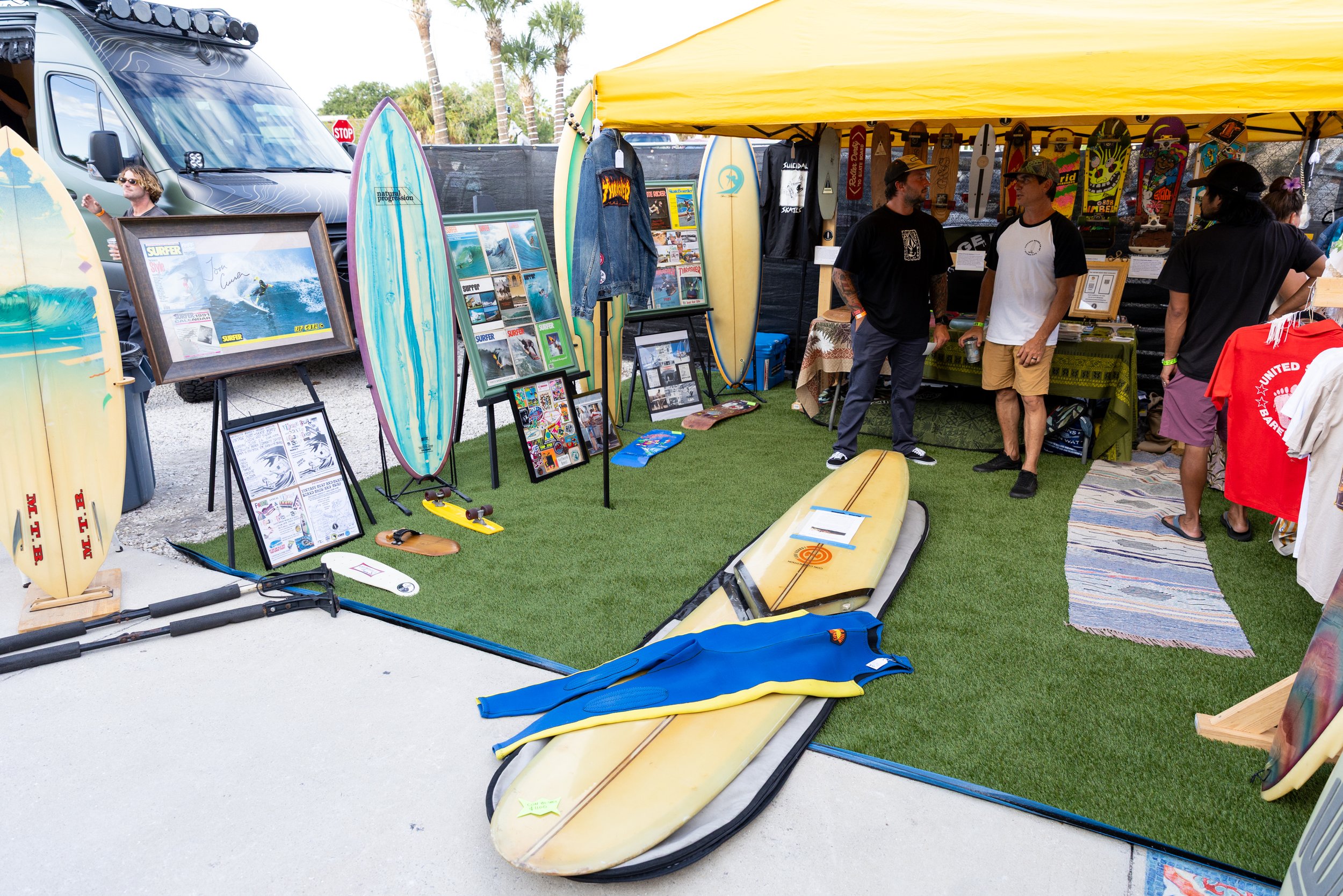 Outdoor display of surfing equipment including surfboards, skateboards, and accessories at a market stall with people browsing and chatting.