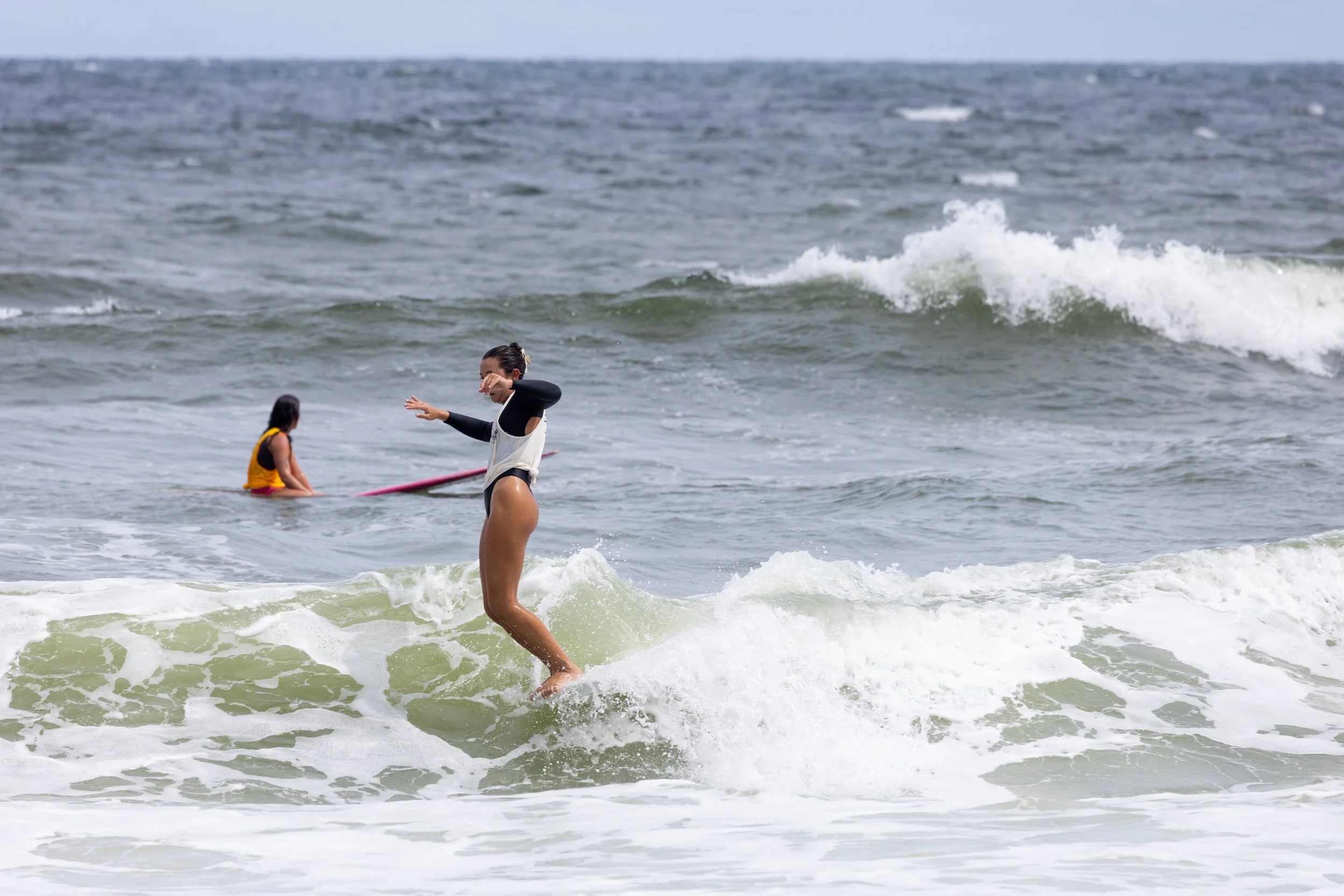 Two women at the beach, one surfing and the other sitting on a surfboard in the water.
