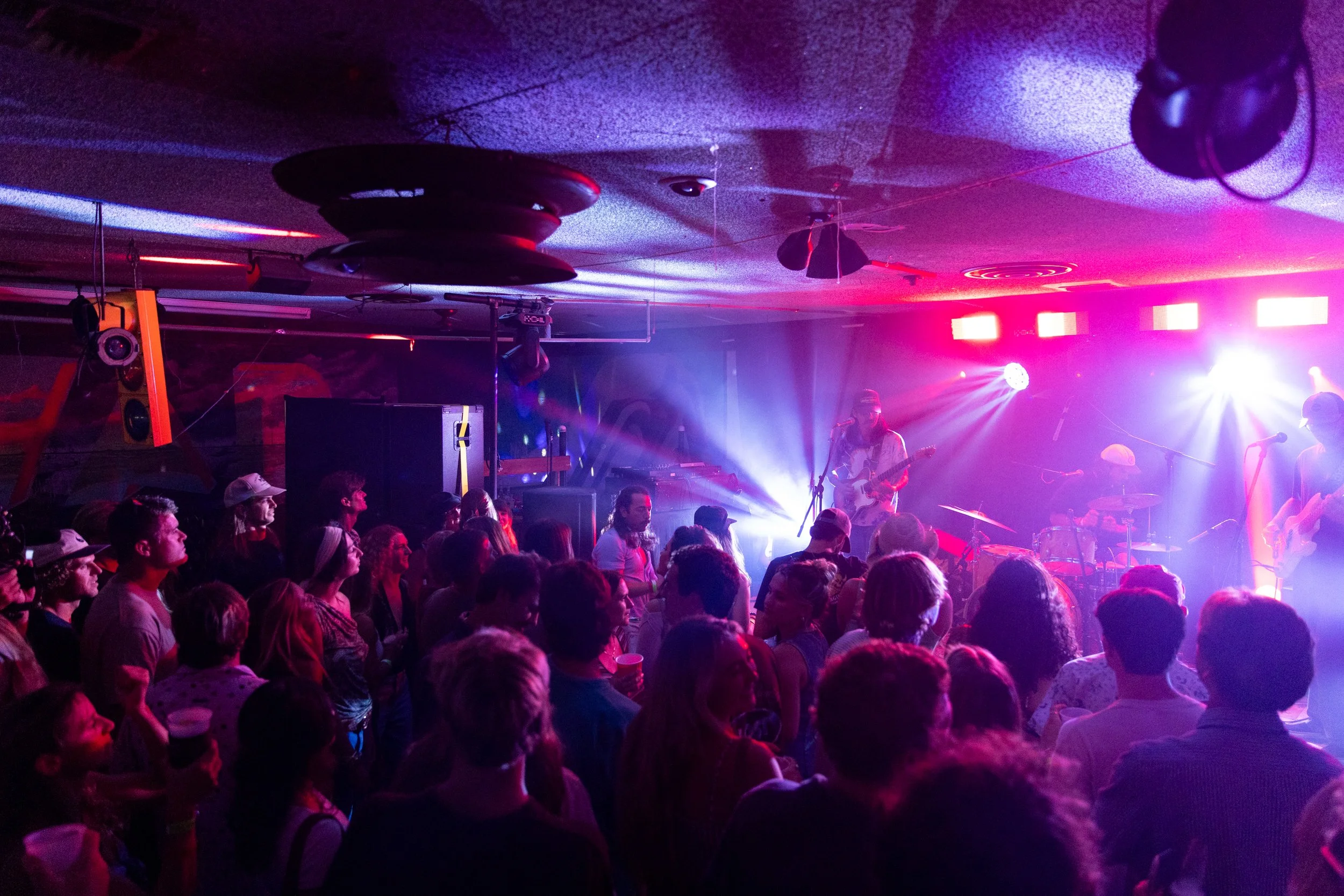 A crowded indoor concert with stage lights, a band performing, and an audience watching.