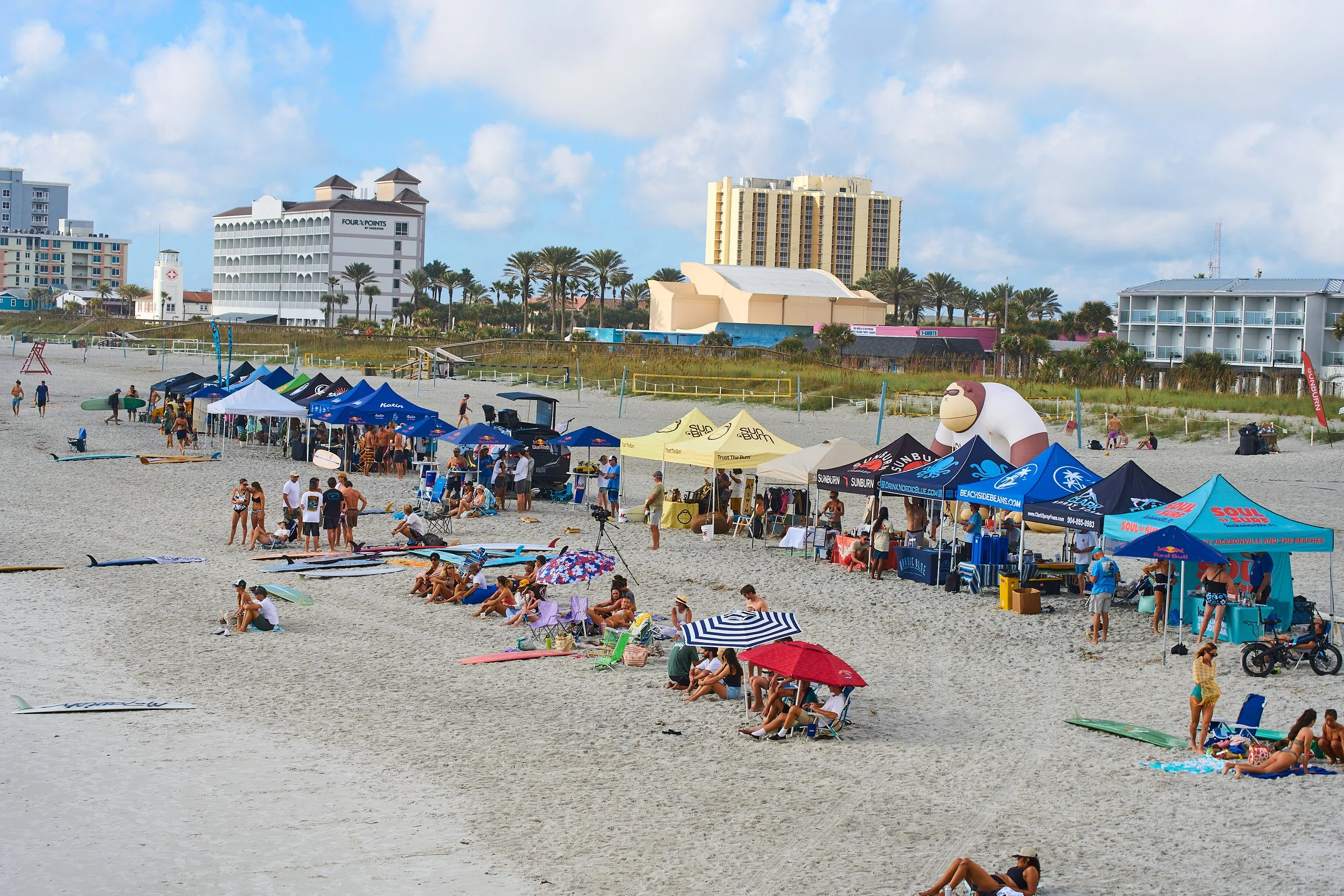 Beach scene with people sitting and standing on the sand, some under umbrellas, and others near tents selling items. There are surfboards lying on the sand, and in the background, high-rise buildings and palm trees are visible against a partly cloudy