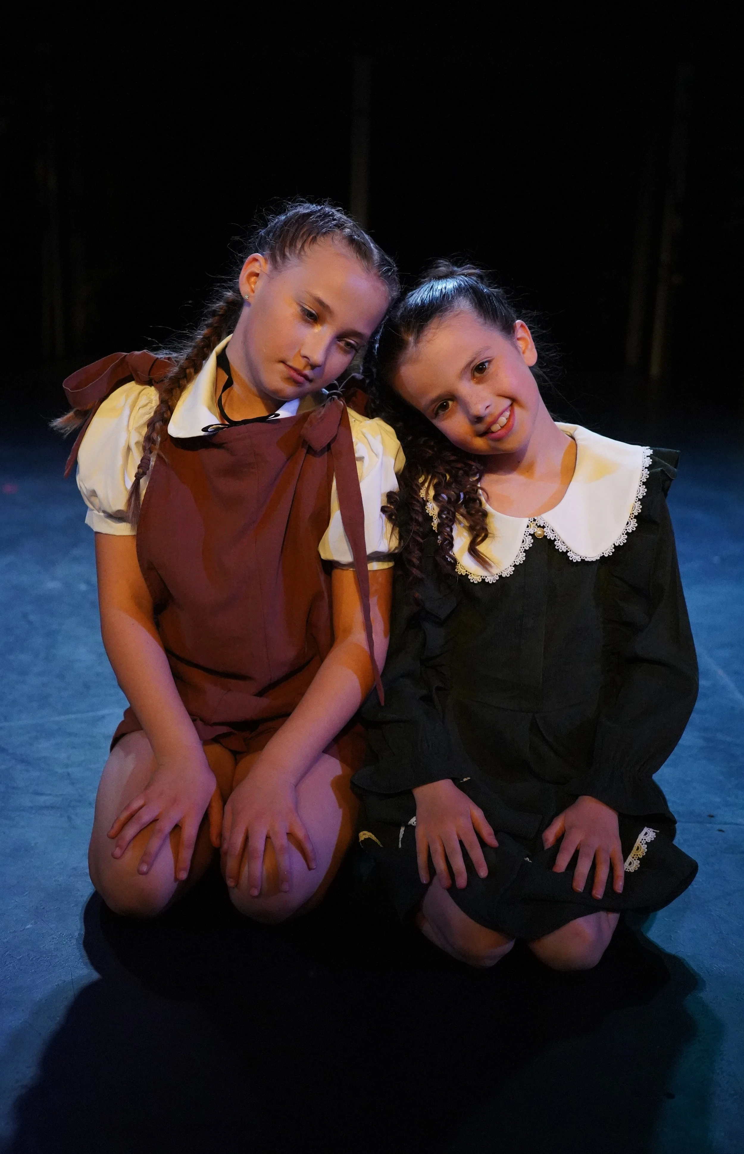 Two young girls in vintage-style costumes sitting on their knees with their hands on their knees, posing on a dark stage.