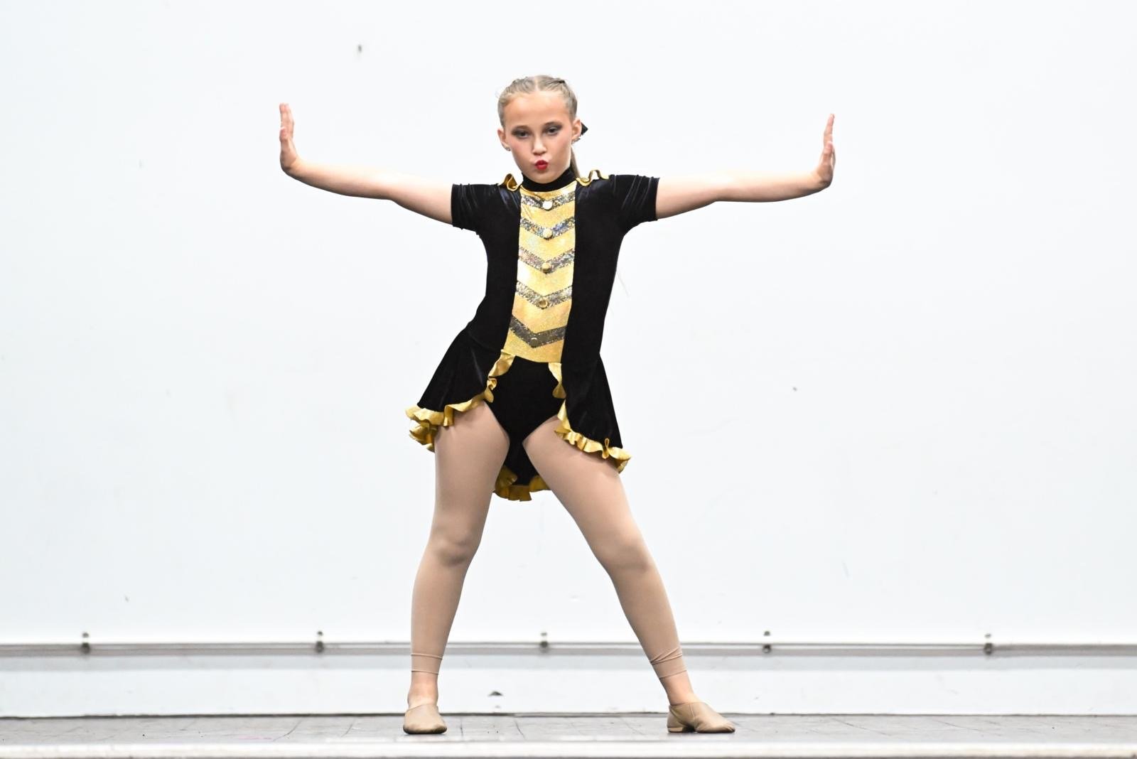 Young girl in a black and gold dance costume performing a dance move with arms outstretched and legs wide apart on stage.