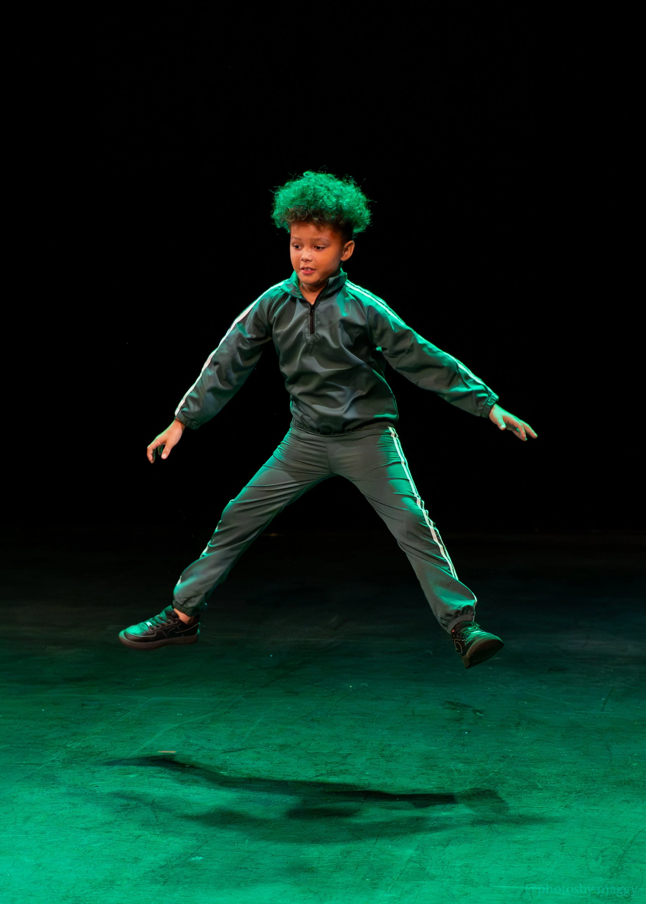 Young boy performing a dance move on stage, wearing a green and gray tracksuit with dark shoes, standing on a dark stage with green lighting.