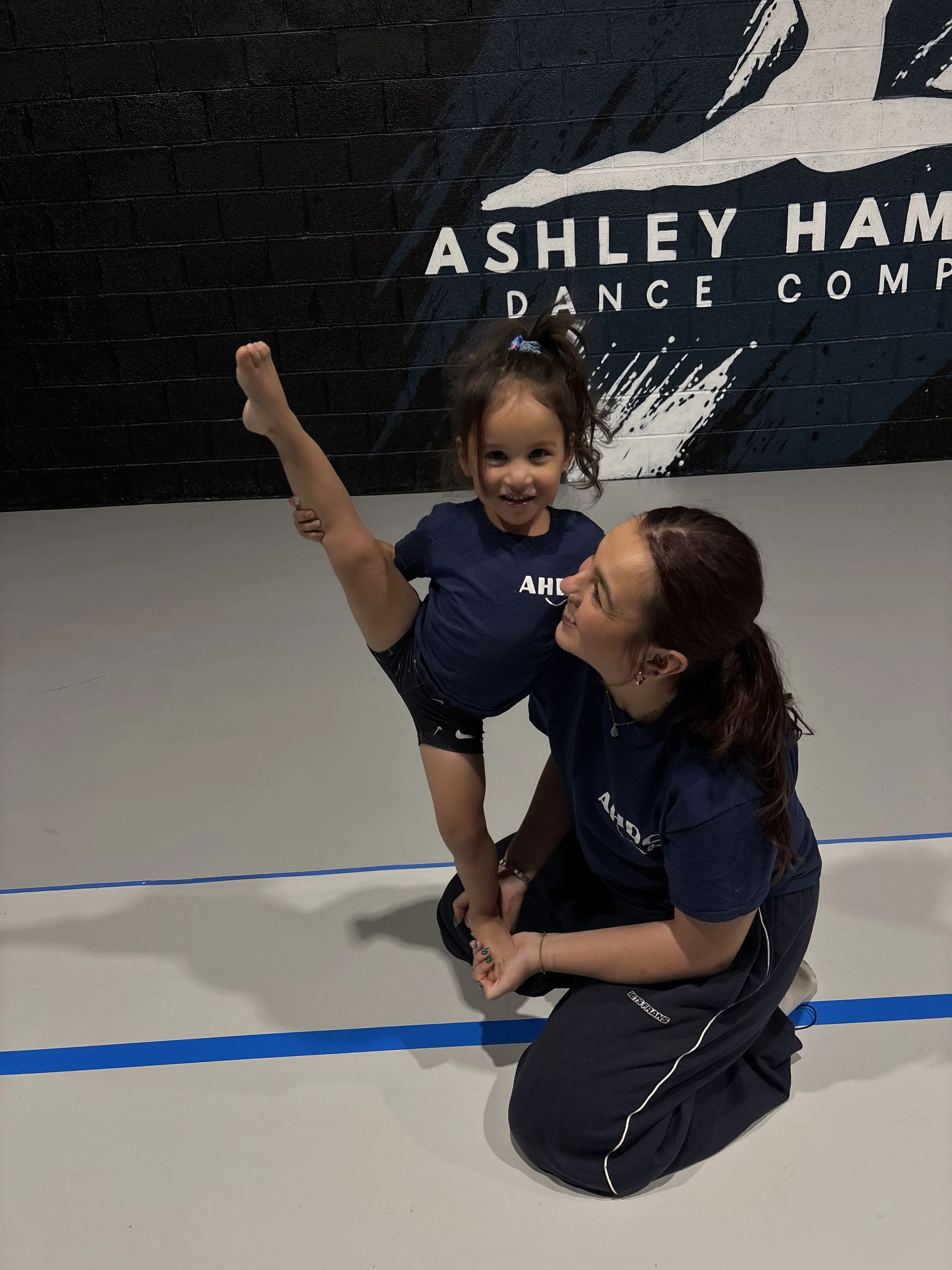 A smiling woman with long brown hair and a ponytail, wearing earrings, is hugging a young girl with red hair in a ponytail. Both are wearing blue shirts. In the background, there are children participating in an activity in a large indoor space with metal walls.
