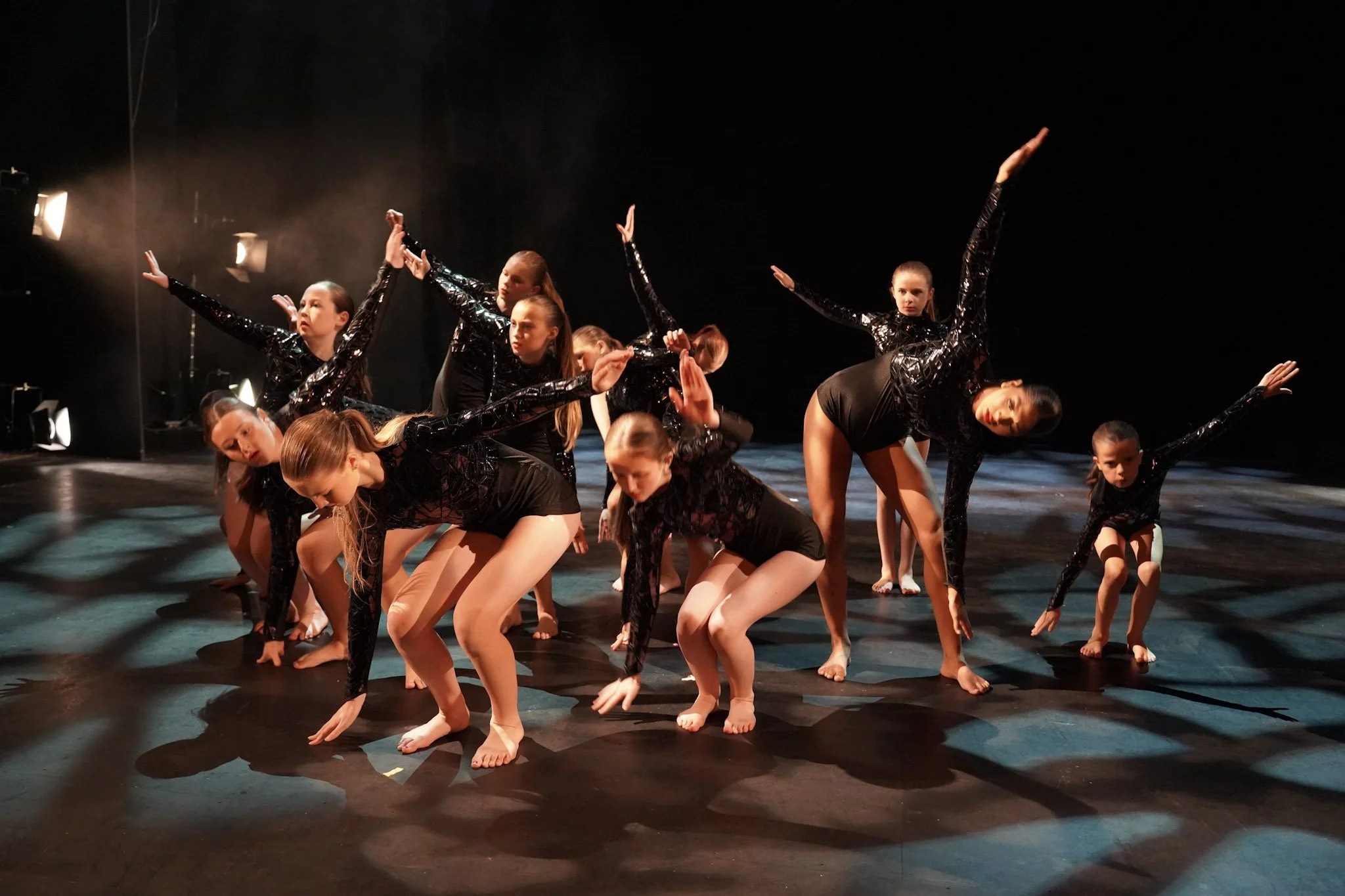 Group of young girls performing a dance on stage, dressed in black costumes with shiny details, with some in poses on the floor and others standing, in a dark theater setting with stage lights.