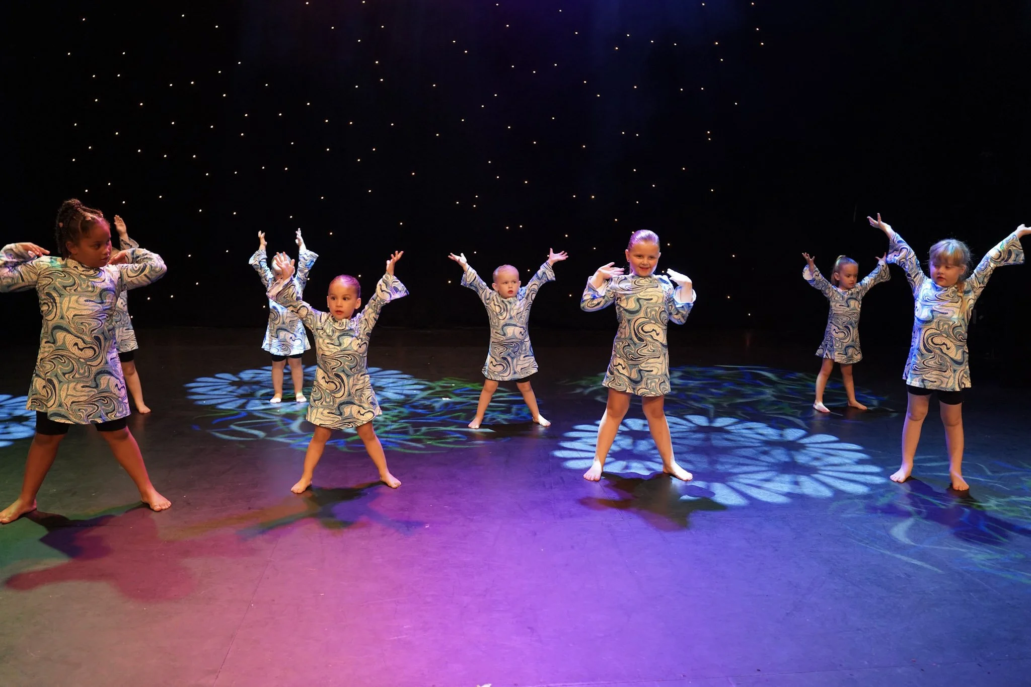 A group of young children performing a dance on stage with a dark background decorated with small bright lights, wearing matching patterned costumes.